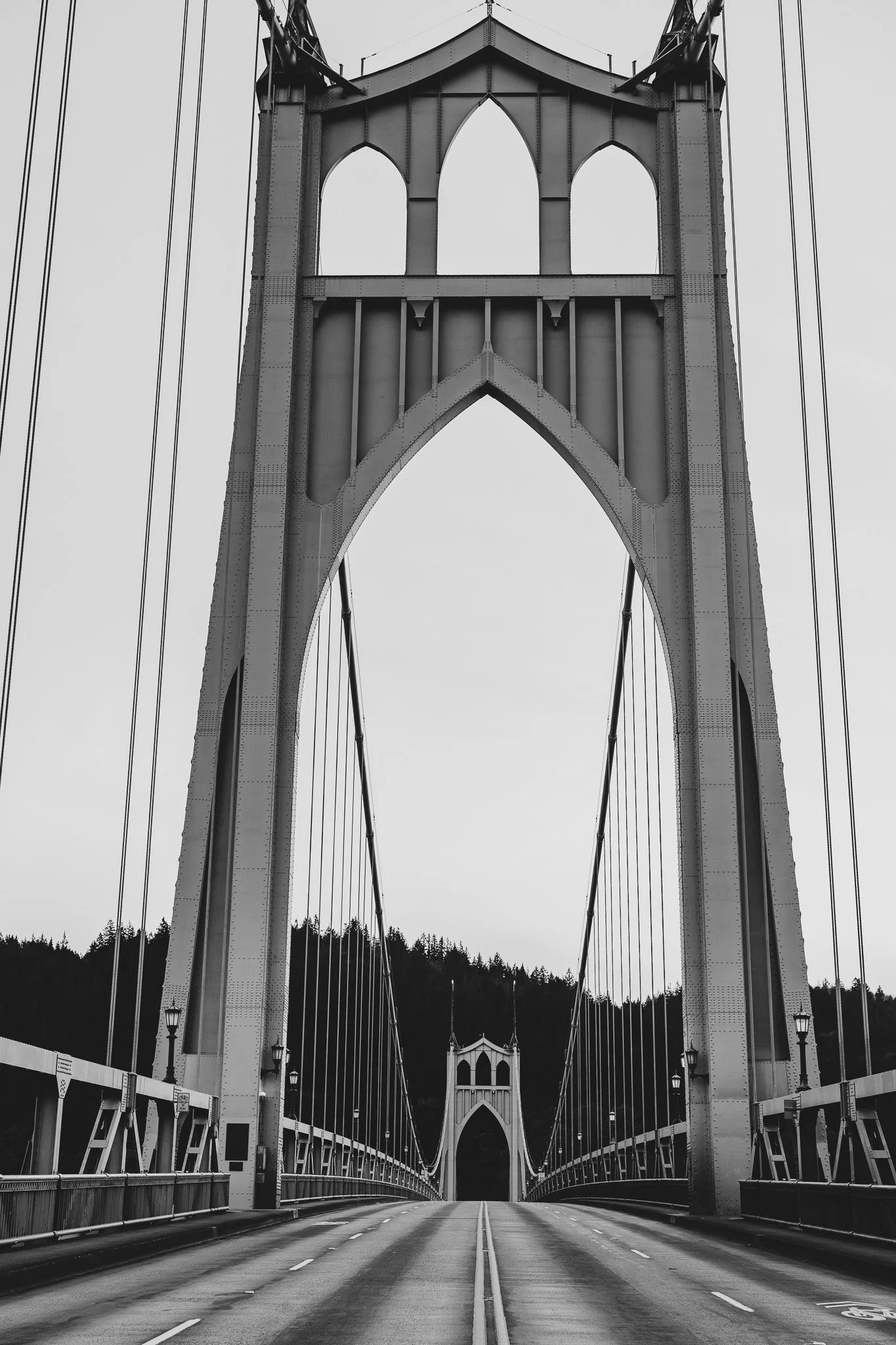 Black and white photo of a suspension bridge with tall towers and cables. The road is empty with no vehicles or people, and there is a forested area in the background beneath the bridge.