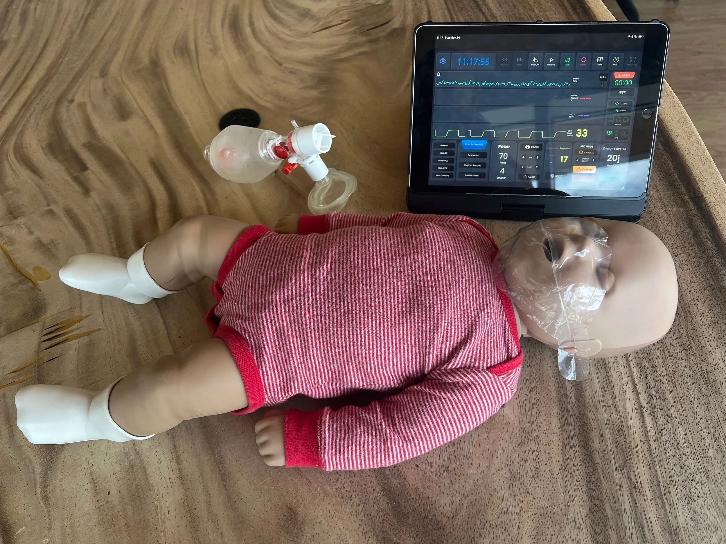 An infant CPR training manikin wearing a red and white striped onesie lies on a wooden table next to an infant-sized bag valve mask resuscitator and a tablet displaying a simulated patient vital sign monitor.