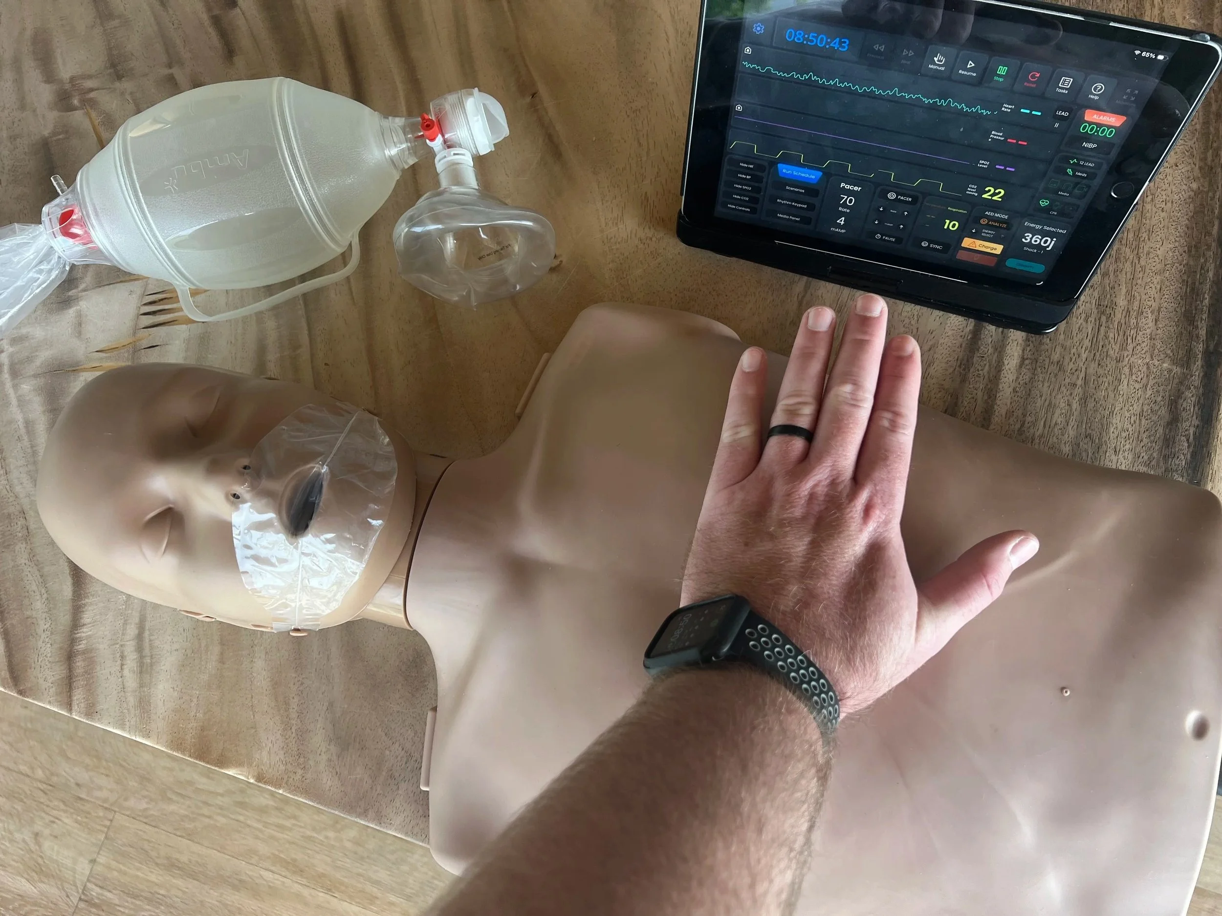 Overhead view of a medical training manikin torso on a wooden table during a CPR or ACLS class. A person’s hand is placed on the chest, next to an Ambu bag ventilation mask and a digital tablet displaying patient monitor vitals.
