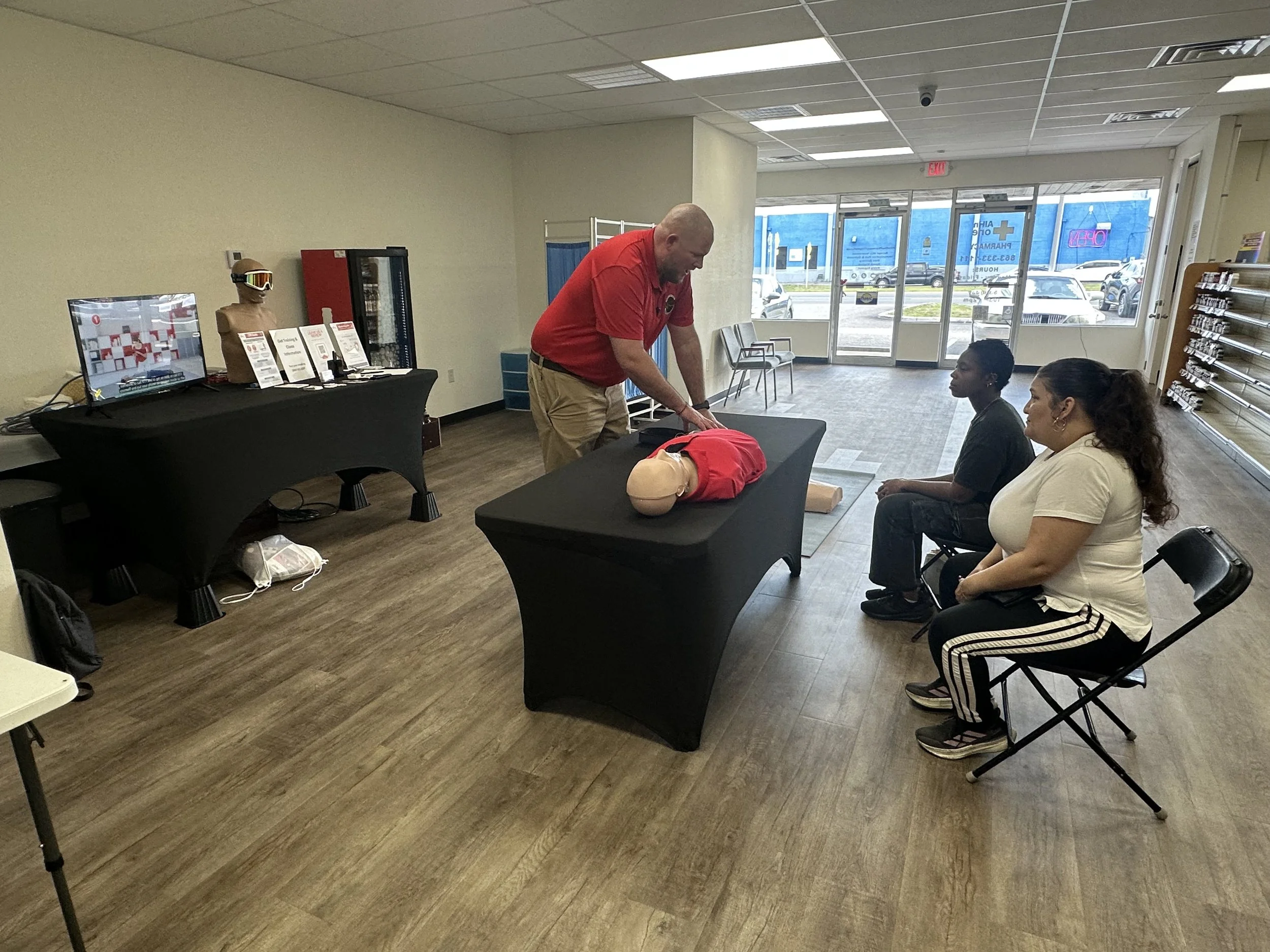 An instructor in a red polo shirt from FACTS First Aid & CPR Training Services demonstrates chest compressions on a CPR manikin during a free community partnership event with All in One Pharmacy, located off Highway 92 in Lakeland, Florida.