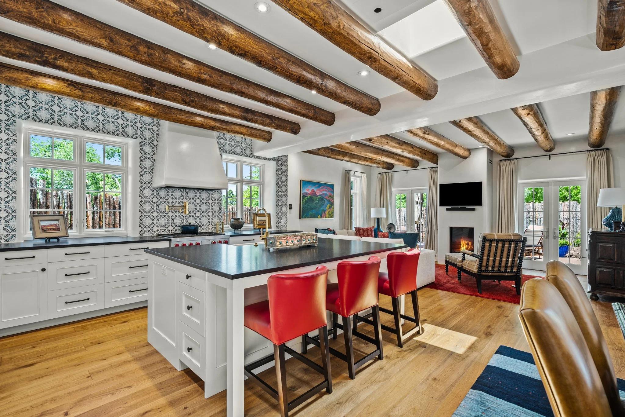 A spacious kitchen and dining area with wooden ceiling beams, white cabinetry, a large island with red stools, and a patterned tile backsplash.