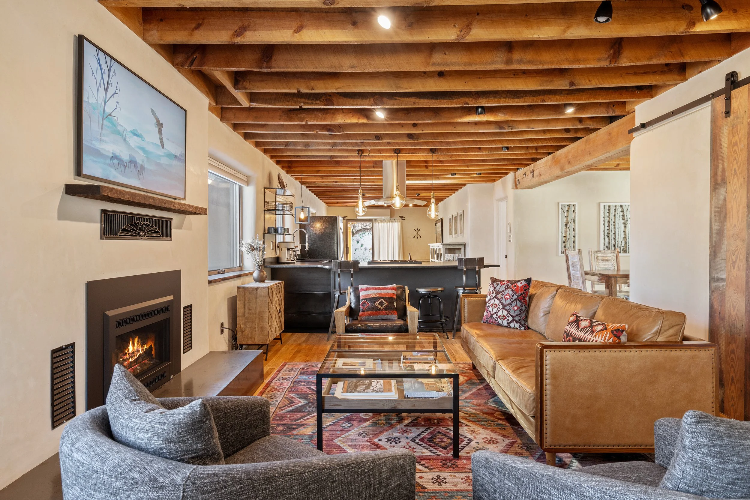 Living room with wooden ceiling, beige sofa with patterned pillows, two blue armchairs, colorful patterned rug, glass coffee table, fireplace, artwork on walls, sliding barn door, open kitchen and dining area in the background.