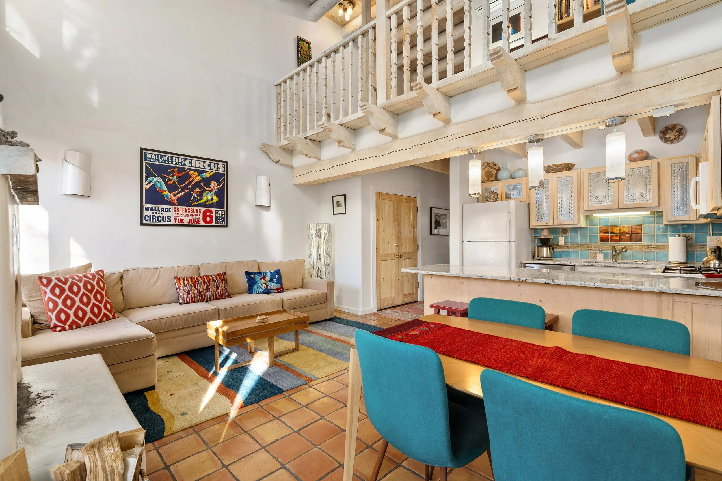 Open-plan living room and kitchen with a beige sectional sofa, colorful throw pillows, a wooden coffee table, a dining table with turquoise chairs, a kitchen counter with flowers, a white refrigerator, and rustic wooden decor.