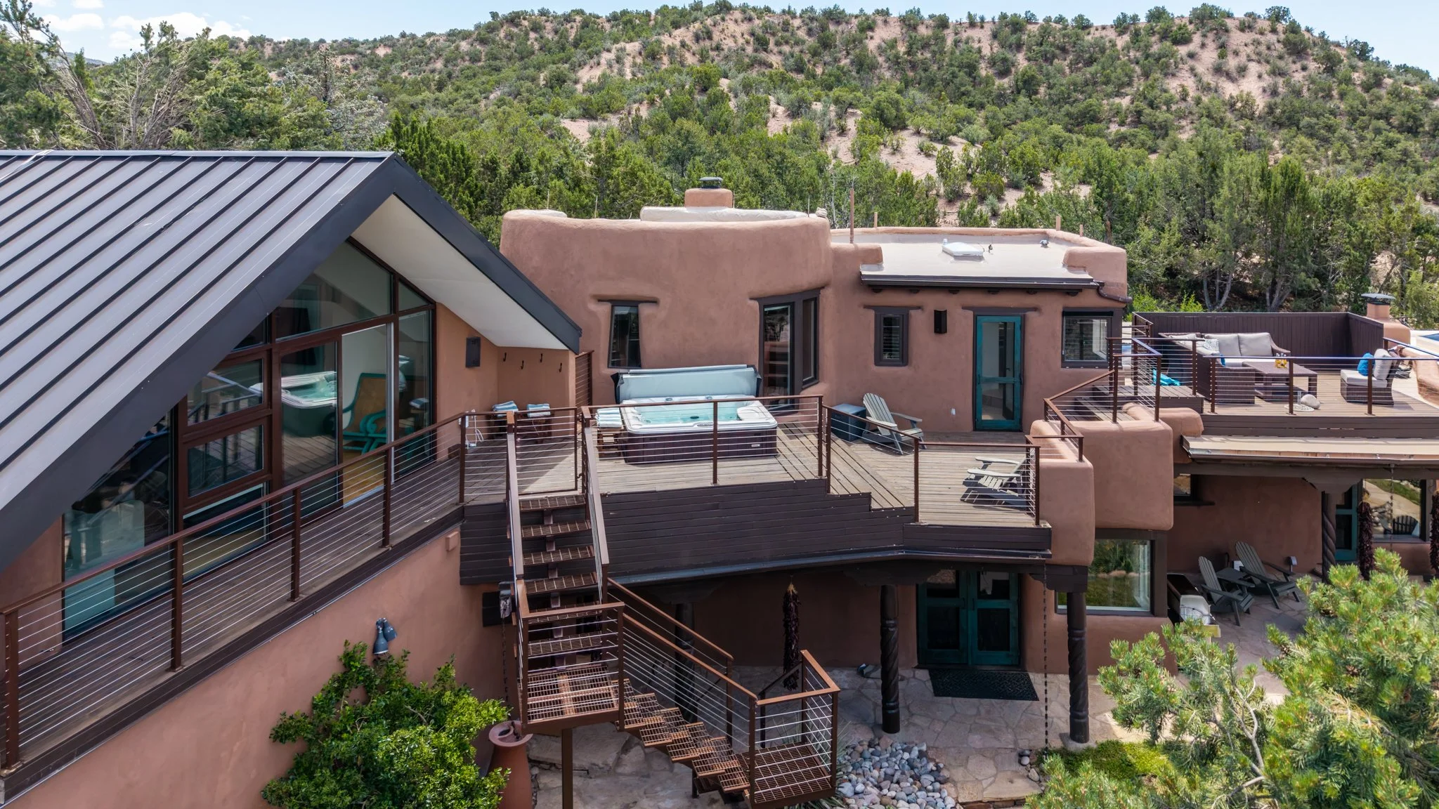 A multi-level house with a rooftop deck, hot tub, and outdoor seating area, surrounded by trees and a hillside.