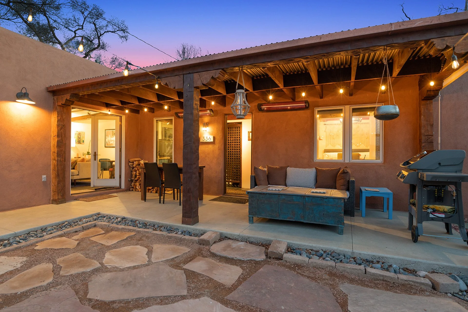 A cozy backyard patio with string lights, a seating area with pillows, a barbecue grill, and a view into the house interior during dusk.