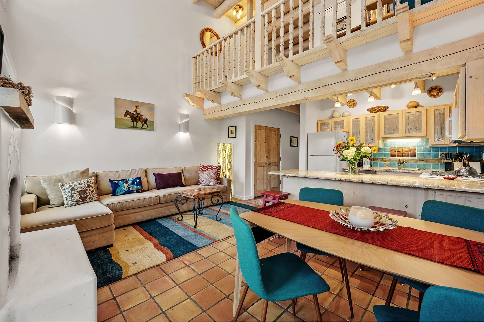 Open-plan living room and kitchen with a beige sectional sofa, colorful throw pillows, a wooden coffee table, a dining table with turquoise chairs, a kitchen counter with flowers, a white refrigerator, and rustic wooden decor.