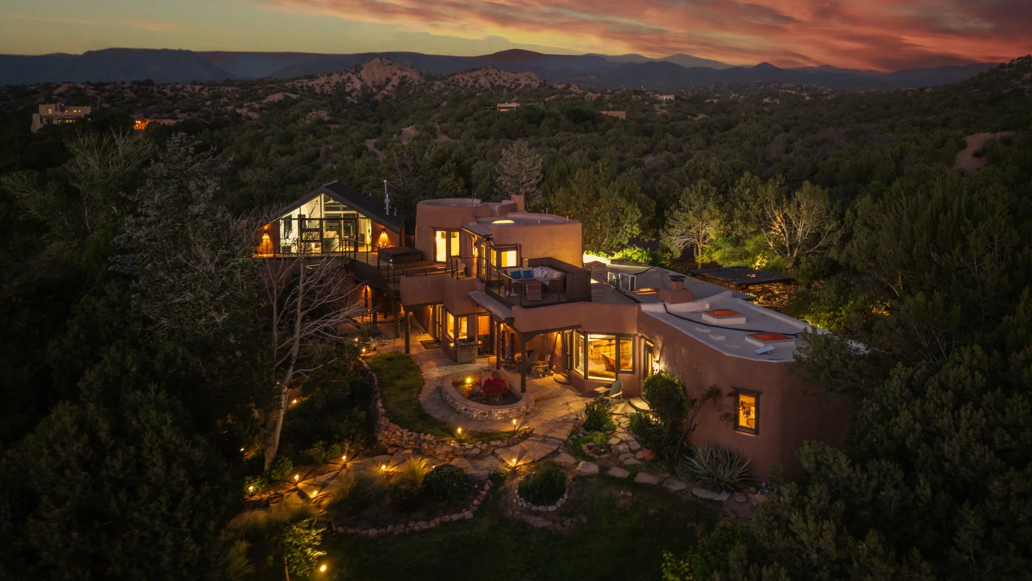 A multi-level house with a rooftop deck, hot tub, and outdoor seating area, surrounded by trees and a hillside.