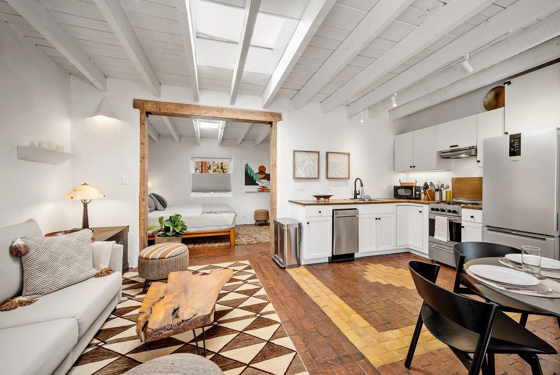 Living room with white walls, exposed ceiling beams, a large white sofa with pillows, a wooden coffee table, a patterned rug, a black dining table with chairs, a wall-mounted TV, a wooden cabinet, and artist paintings on the wall.