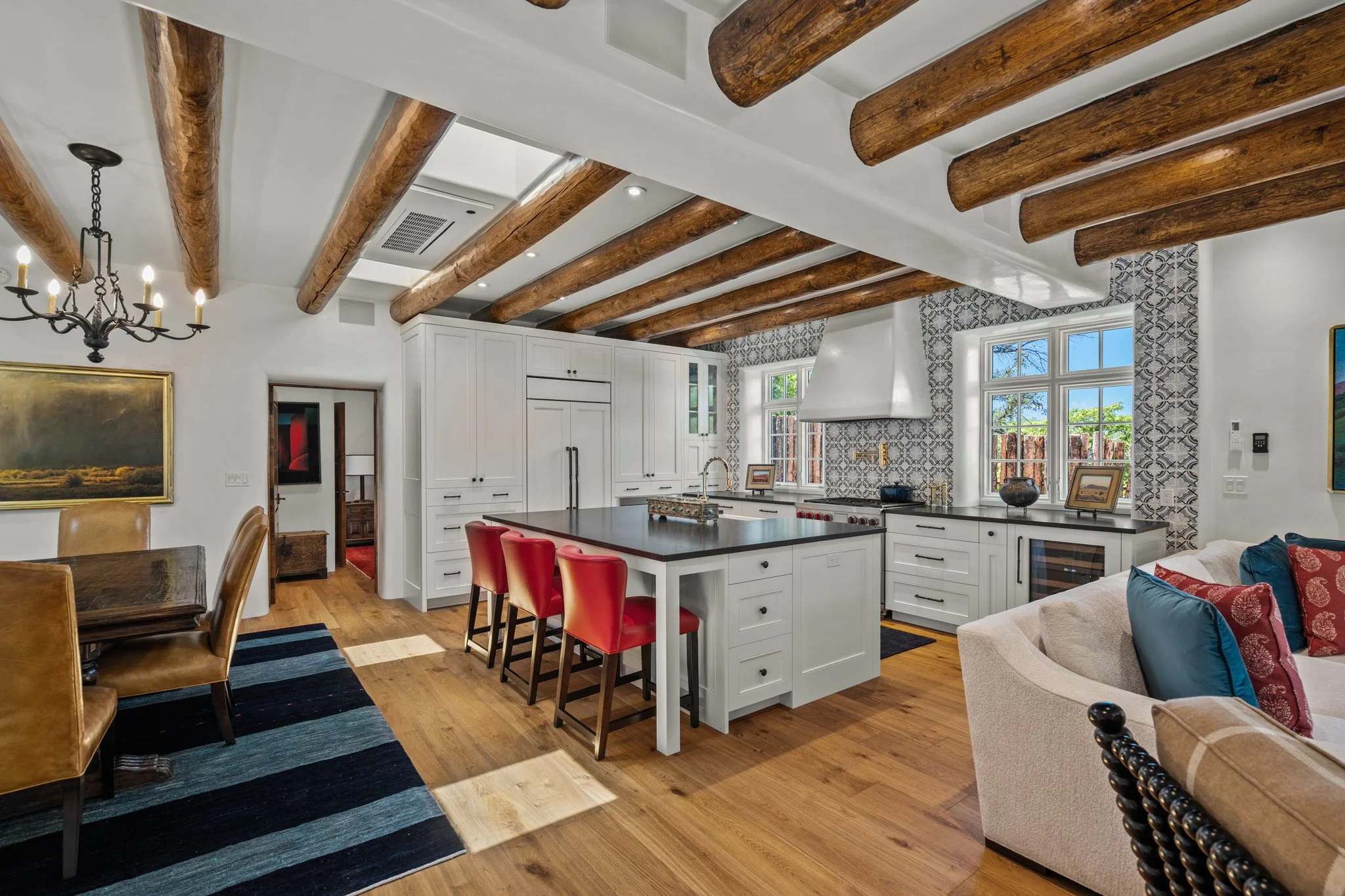 A spacious kitchen and dining area with wooden ceiling beams, white cabinetry, a large island with red stools, and a patterned tile backsplash.