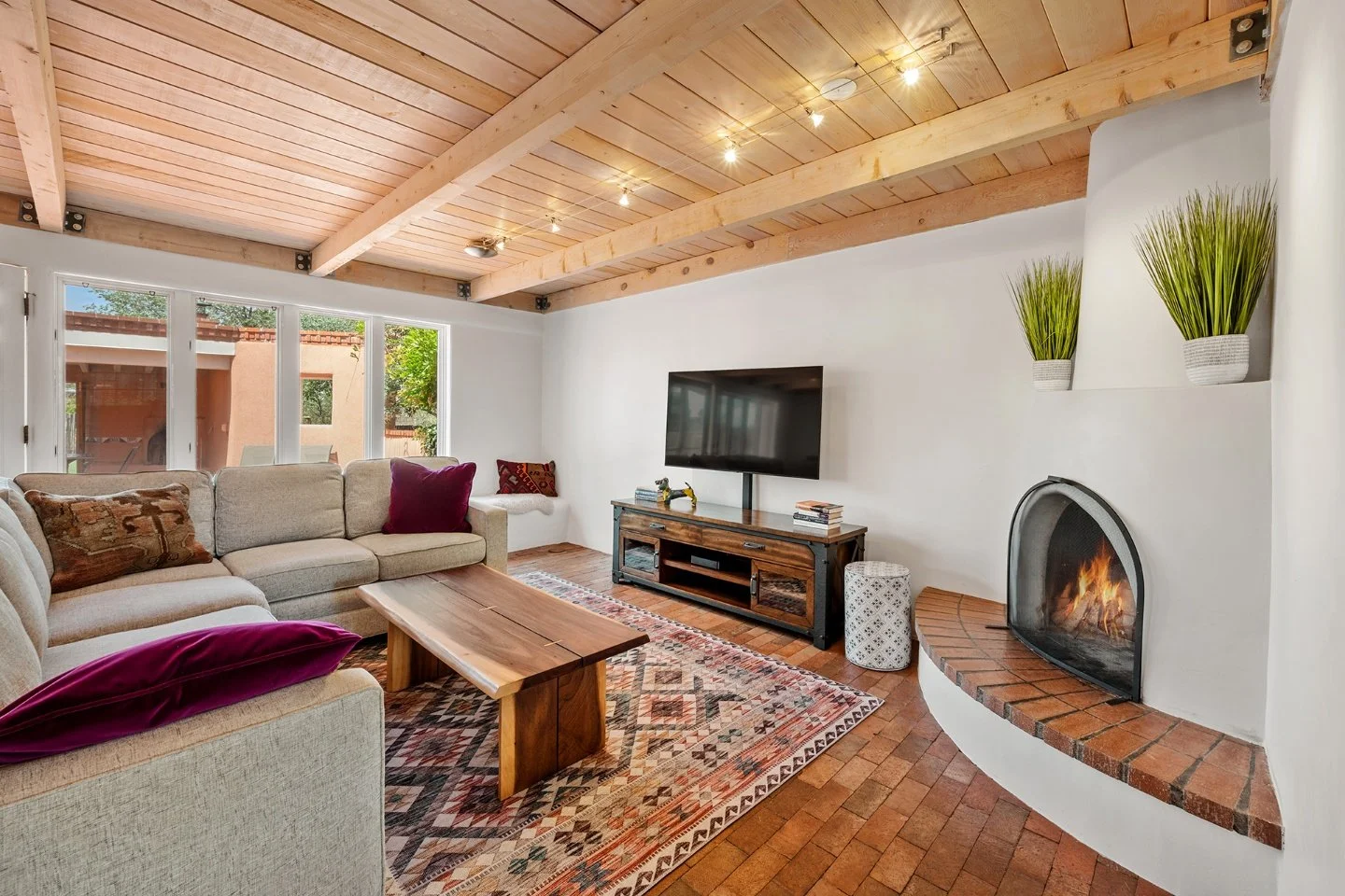Living room with a wooden ceiling, a beige sectional sofa with colorful pillows, a wooden coffee table, a patterned rug, a TV mounted on a white wall, a corner fireplace with brick hearth, and large windows showing trees outside.