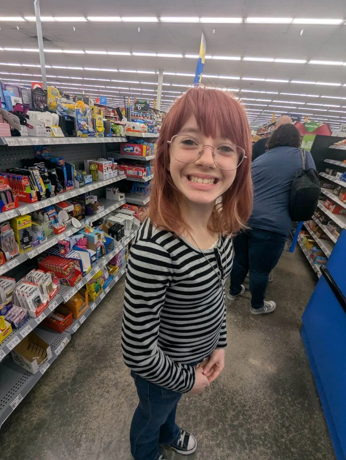 A smiling girl with red hair and glasses in a store aisle, wearing a black and white striped long-sleeve shirt and jeans.
