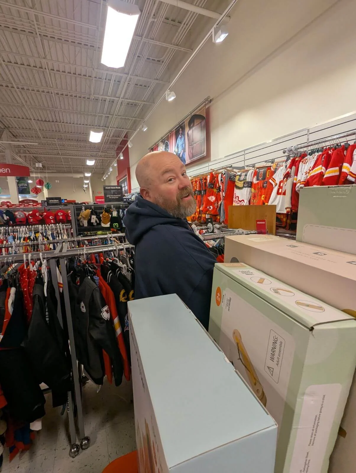 A man with a beard and bald head shopping in a store, surrounded by sports jerseys and boxed holiday lights, looking back at the camera.