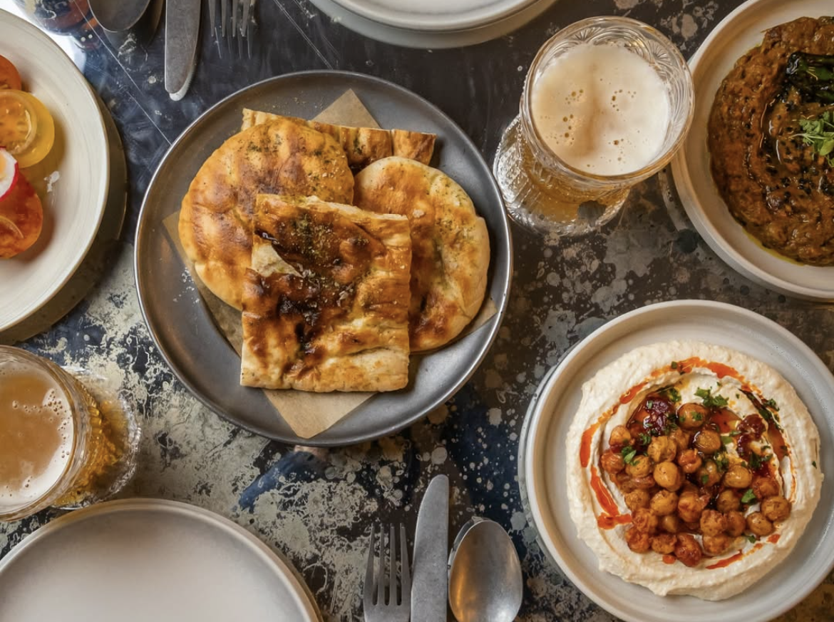 A table of Persian dishes including a plate of flatbread, chickpea hummus, fresh salads and Persian drinks.