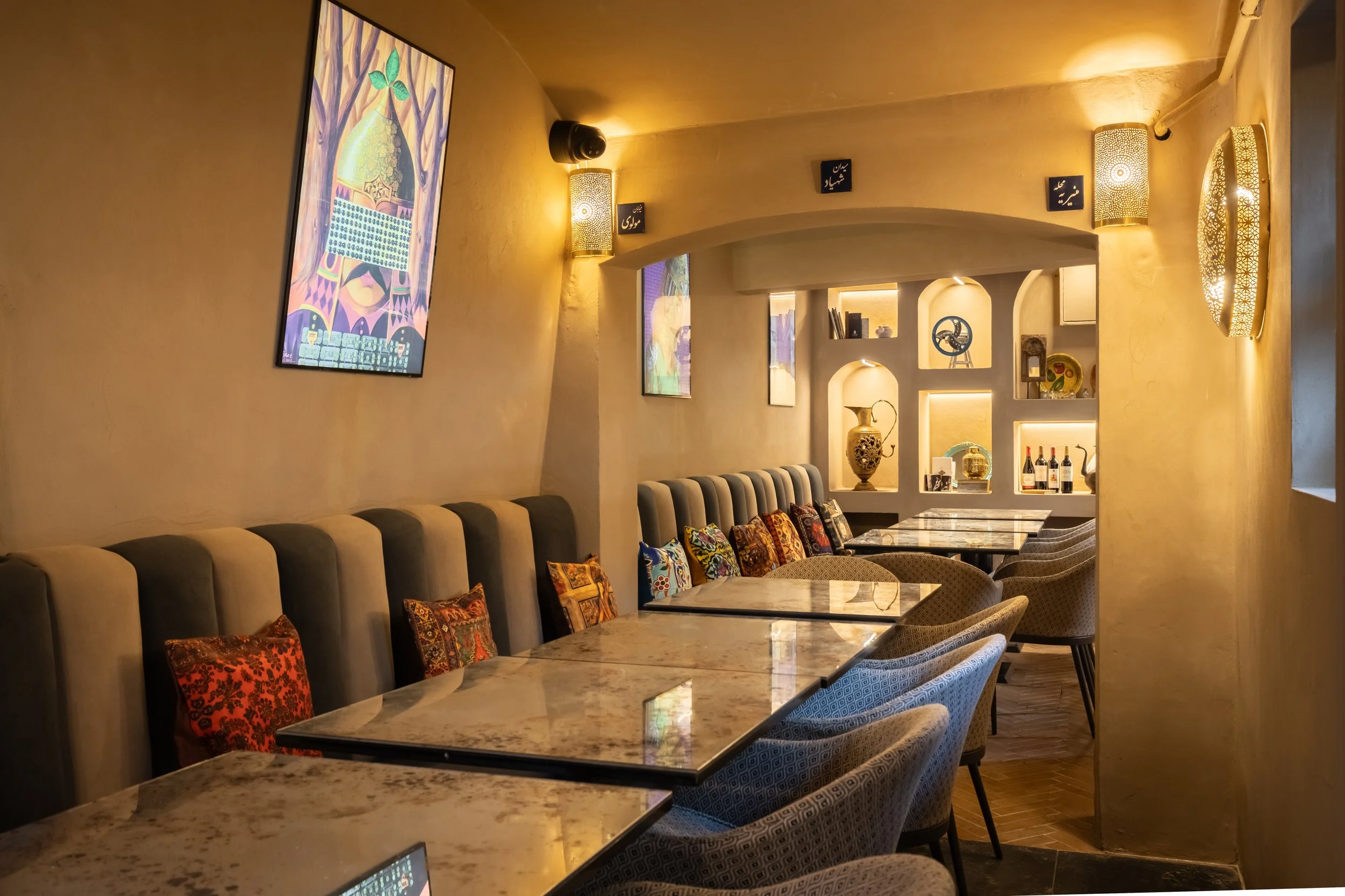 Interior view of a Ember By Chelo restaurant in Maida Vale London with marble tables, cushioned seating, colourful decorative pillows, warm lighting, wall art, and display shelves with decorative items and bottles.