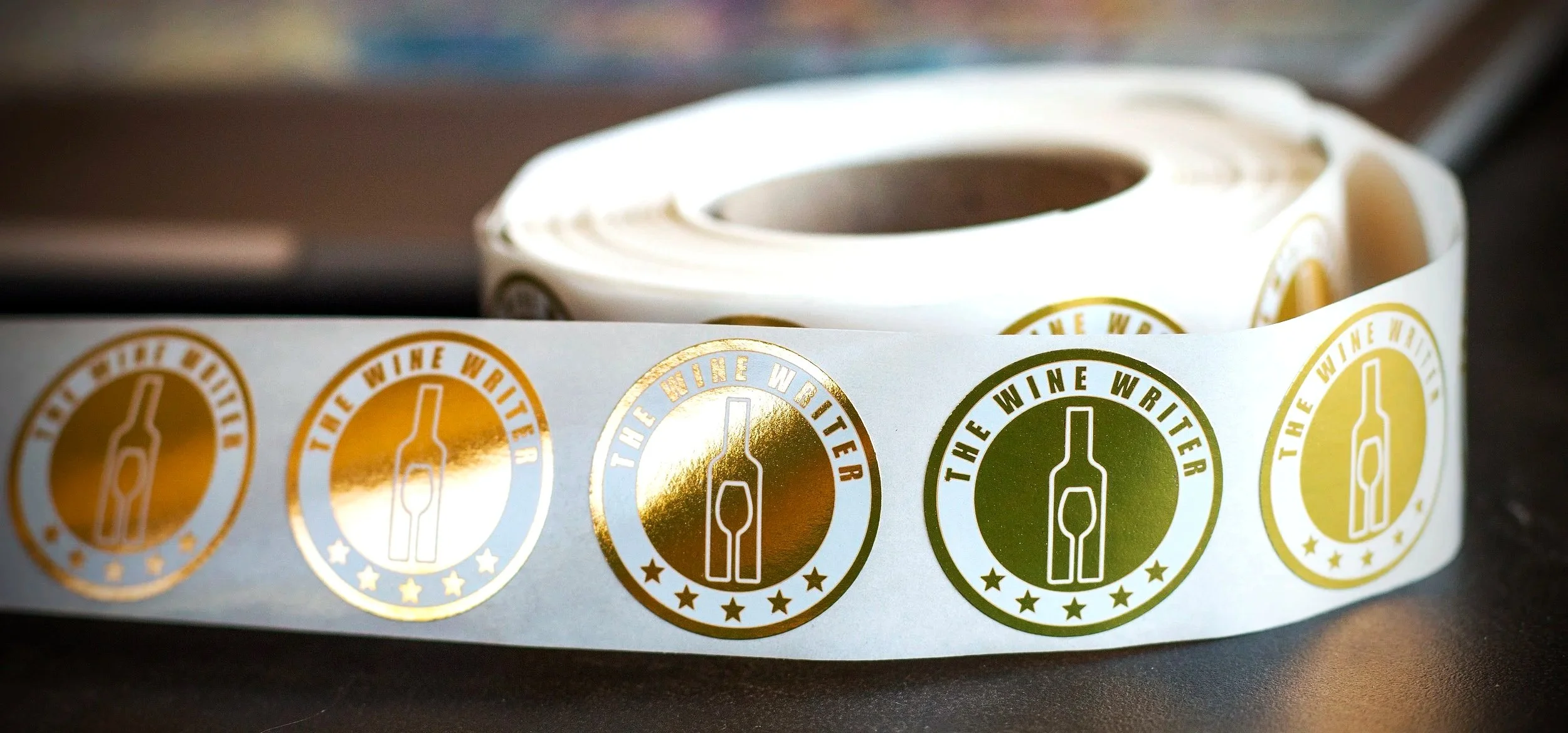 Roll of stickers with gold and green foil labels reading 'The Wine Writer' with a wine bottle and glass icon, partially unrolled on a dark surface.