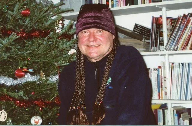 A scientist, Dr. Hans Peterson, is wearing braids gifted by Yellow Quill First Nation. He is next to a Christmas tree and is also wearing a burgundy toque.