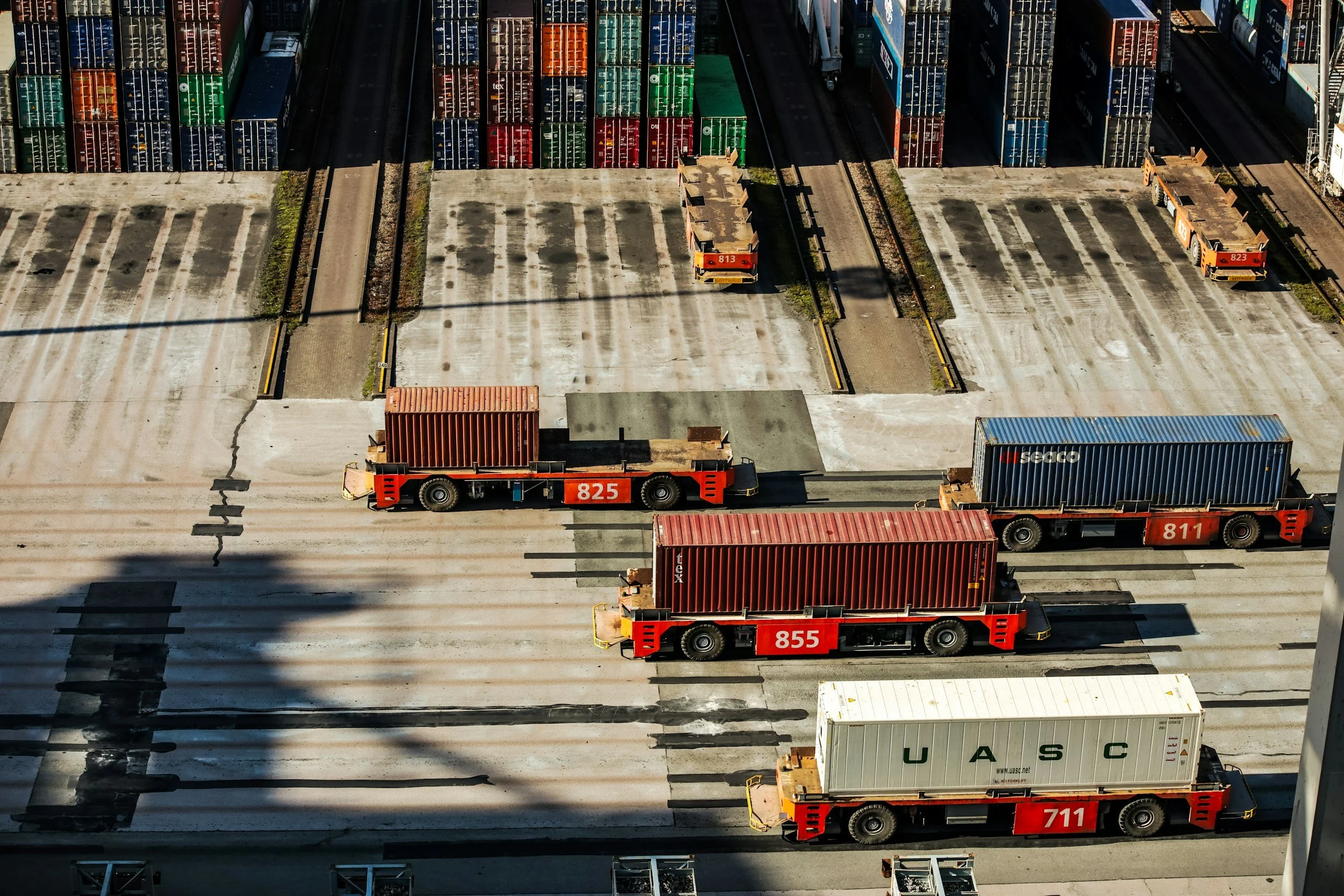 Vista aérea de un deposito de contenedores y vehículos de carga en un puerto, con camiones de transporte de contenedores de colores y contenedores metálicos apilados.