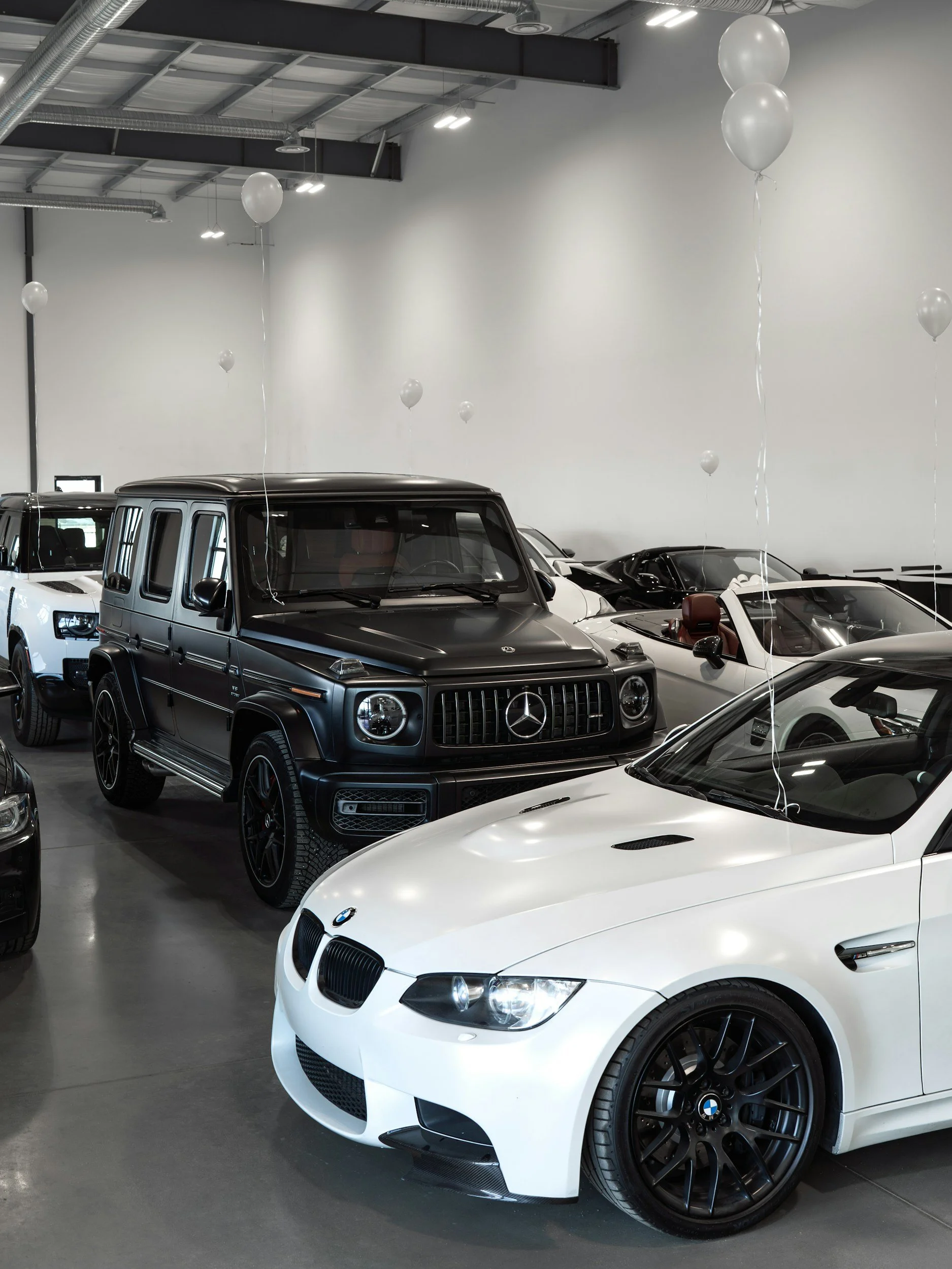 Varios autos de lujo en un showroom, incluyendo un BMW y un Mercedes-Benz, con globos blancos decorativos suspendidos del techo.