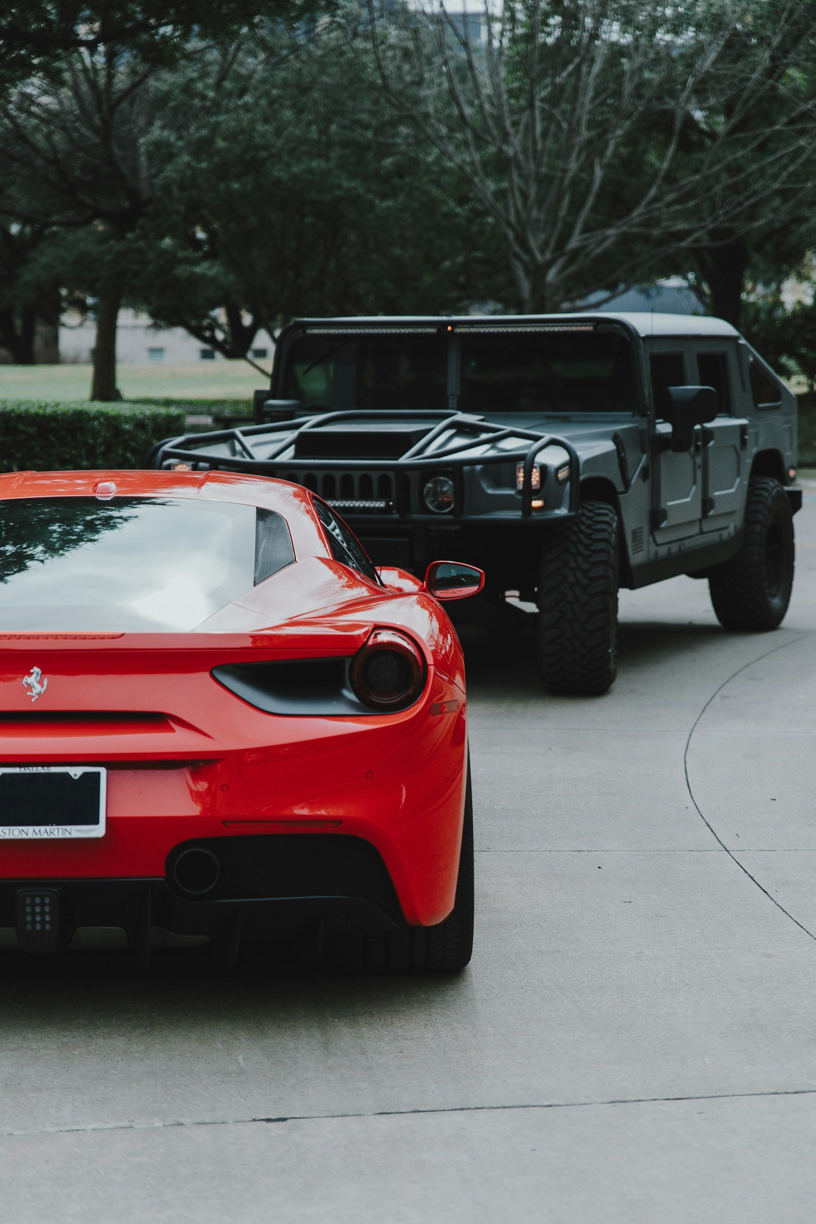 Un automóvil deportivo rojo, una Ferrari, en primer plano y parcialmente visible, y un vehículo todoterreno negro en segundo plano, con árboles verdes y césped en un parque o calle en un día nublado.