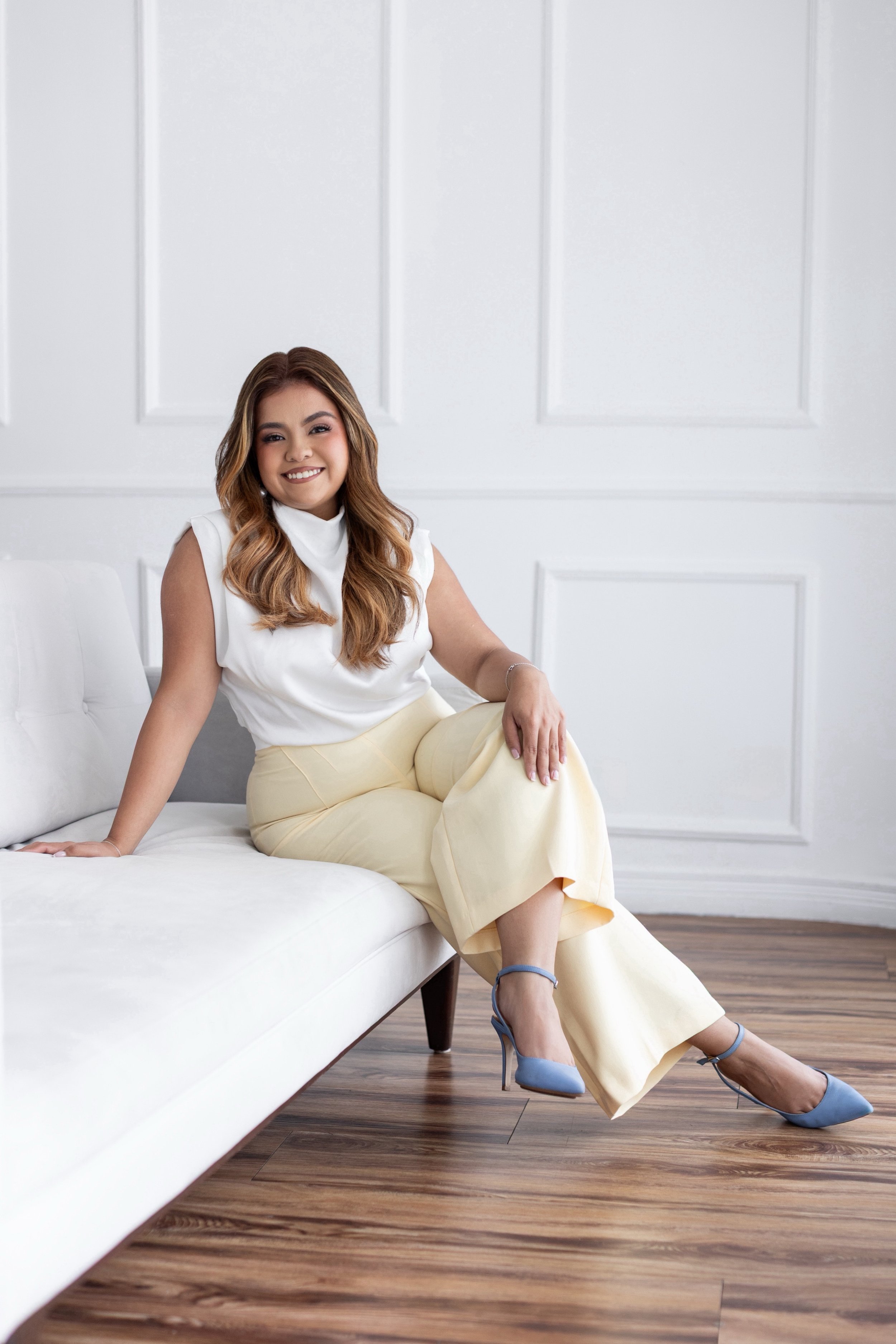 Mujer sonriente sentada en un sofá blanco en una sala con paredes blancas y pisos de madera, vestida con blusa blanca y pantalones beige, usando tacones azules.