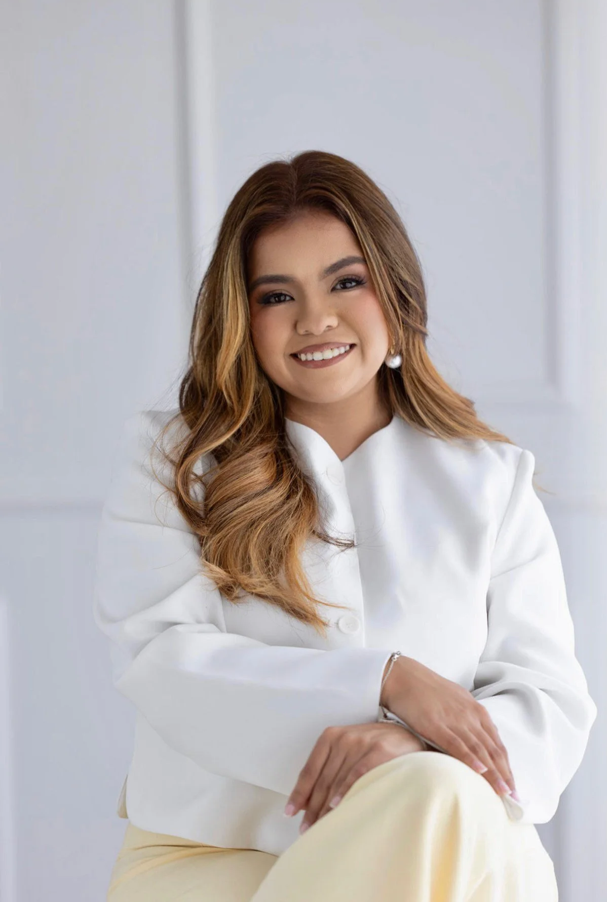 Retrato de una mujer sonriendo, con cabello castaño, vestida con blusa blanca y pantalones claros, en un ambiente interior blanco y luminoso.