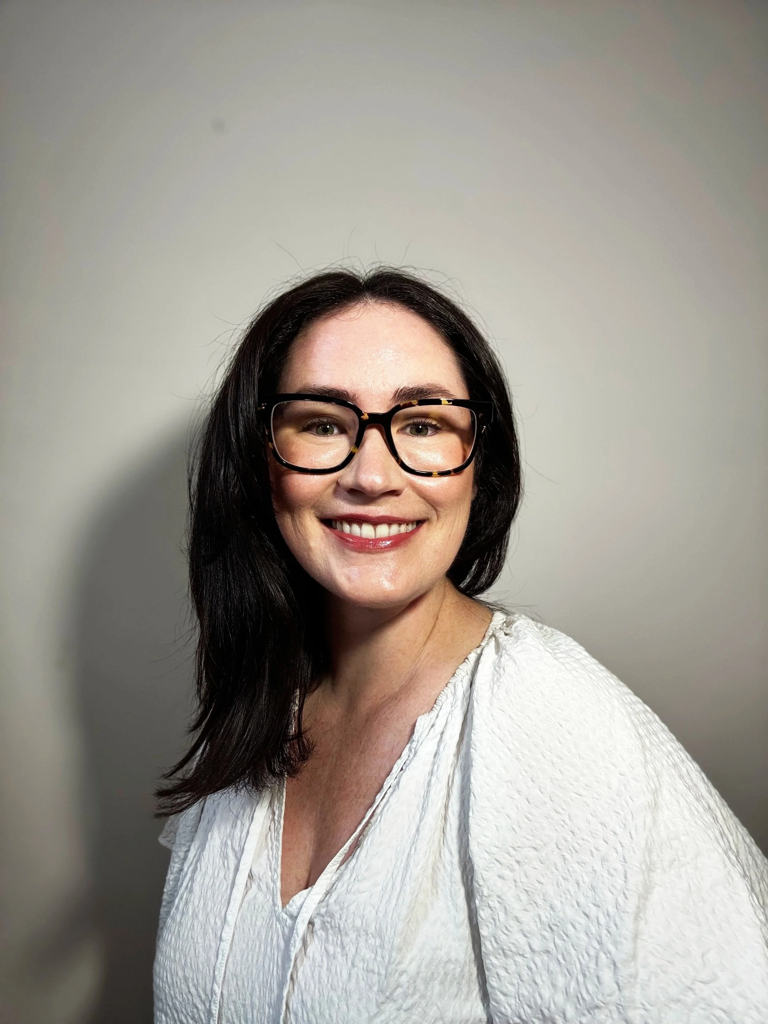 A woman with long dark hair, wearing glasses and a white textured blouse, smiling in front of a plain light gray wall.