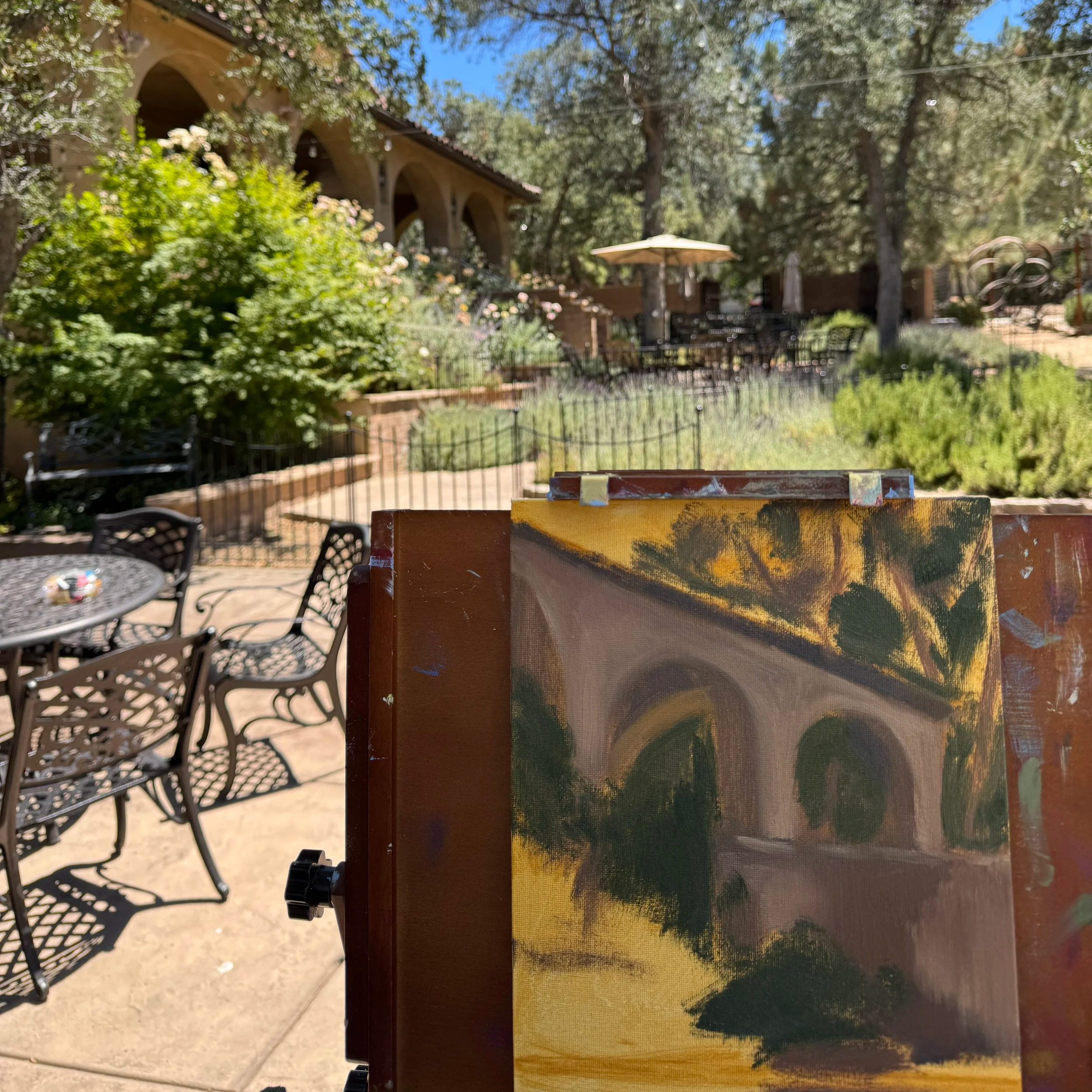 A colorful painting of a landscape with green, black, and yellow hues leaning against a surface in an outdoor setting. In the background, outdoor furniture, trees, and a blue sky are visible.