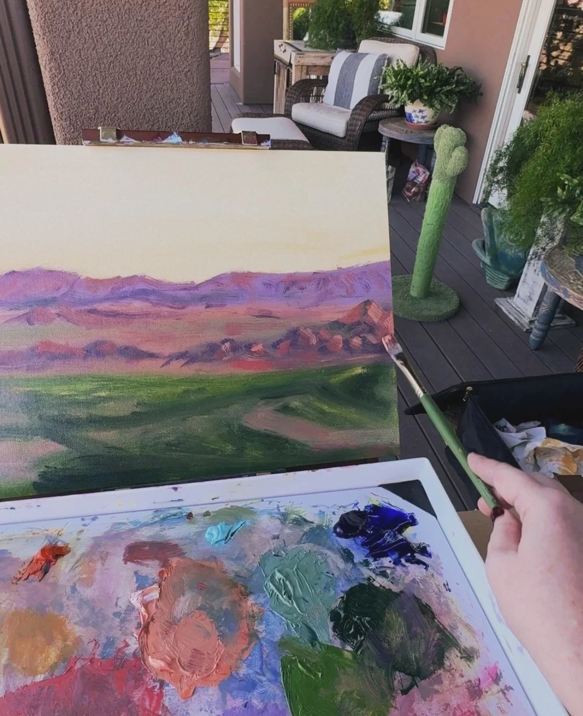 A person painting an outdoor landscape scene on a canvas, with visible brushstrokes of mountains, sky, and grass, using a palette with various colors, on a balcony with chairs, plants, and tables in the background.
