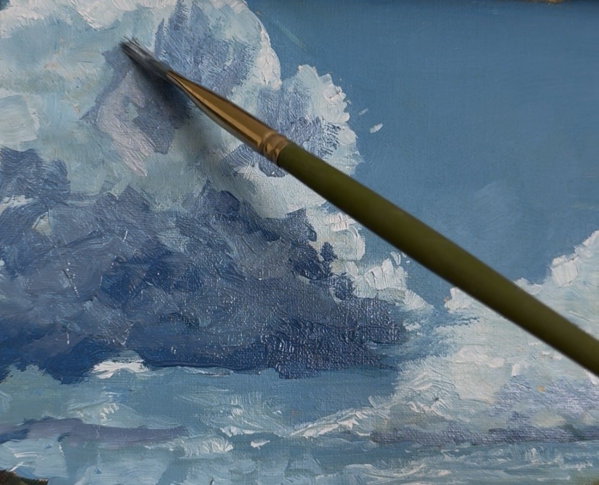 Close-up of an artist's brush painting a cloudy sky on a canvas with shades of blue and white.