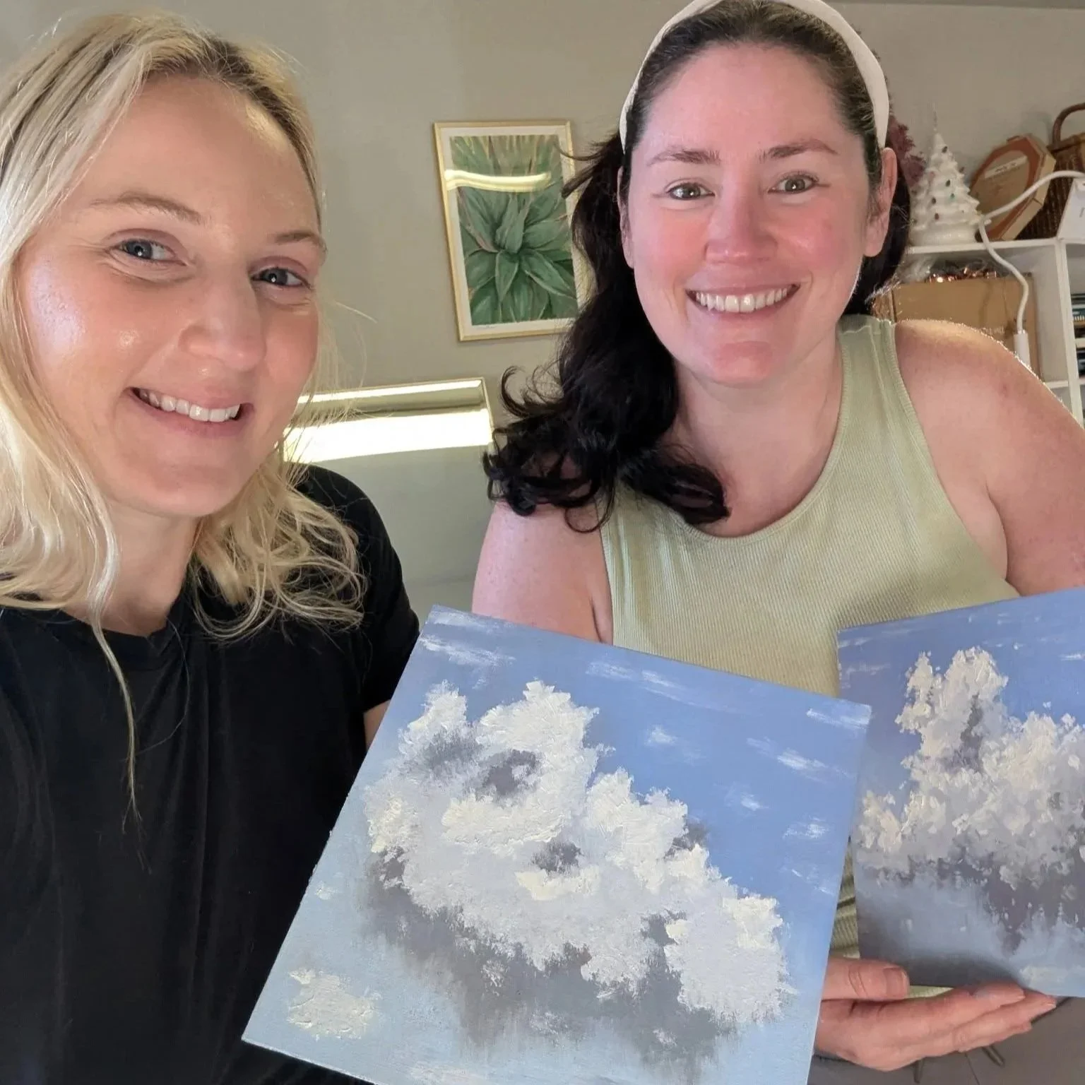 Two women smiling, one with blonde hair and the other with dark hair, holding paintings of clouds in the sky, indoors with a framed plant picture on the wall in the background.