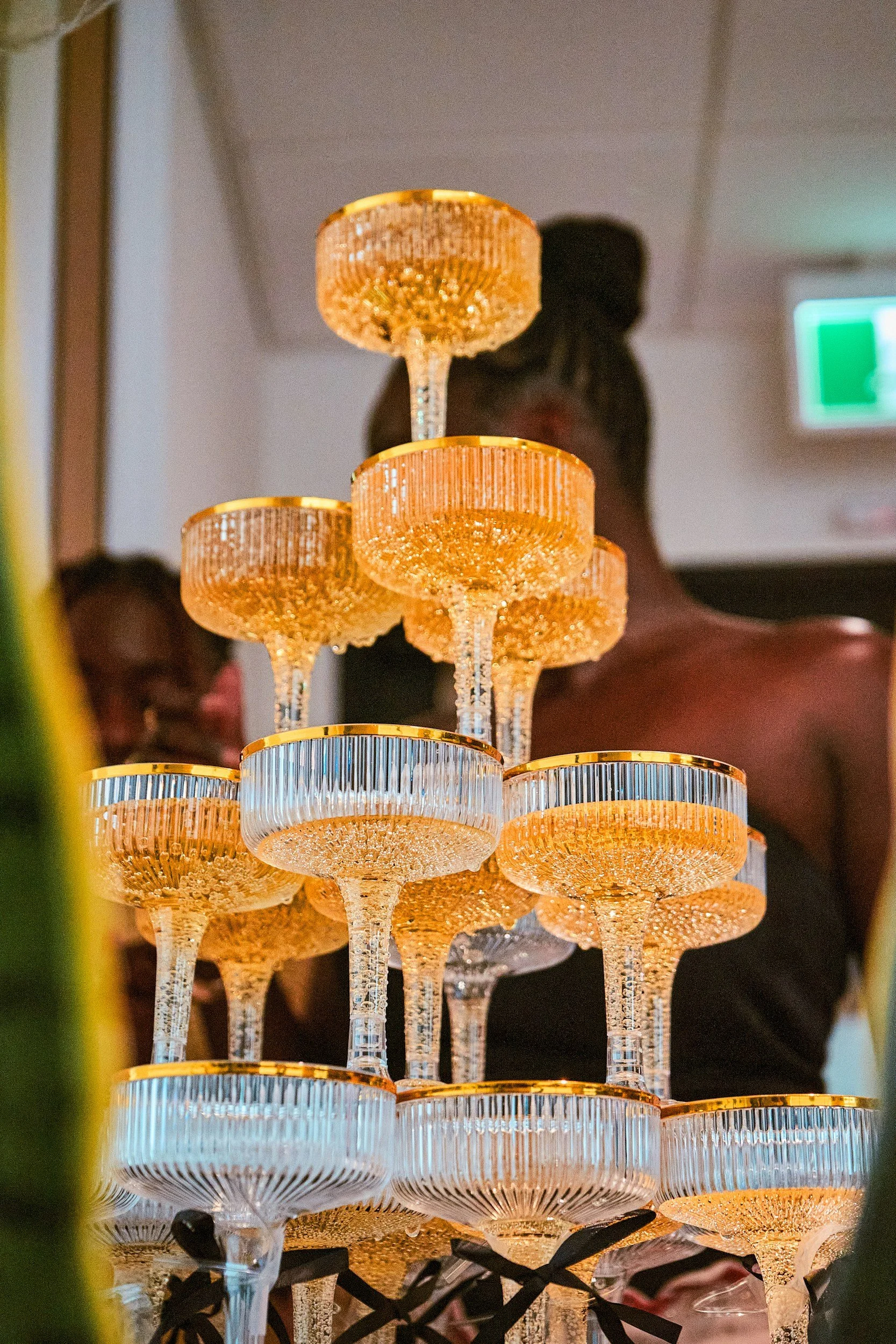 A pyramid of champagne glasses with gold rims, stacked in a layered chandelier-like arrangement in an indoor setting, with a blurred background featuring a person and a green exit sign.