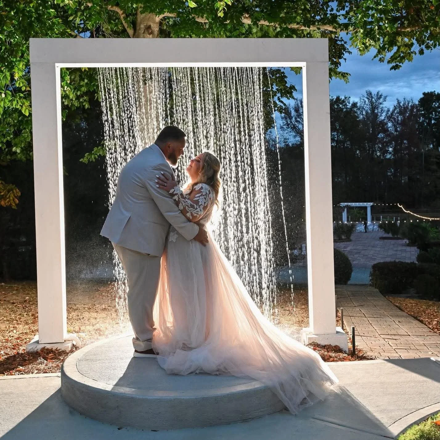 A bride and groom sharing a romantic moment under a small waterfall feature at dusk, with the bride in a flowing pink gown and the groom in a light suit.