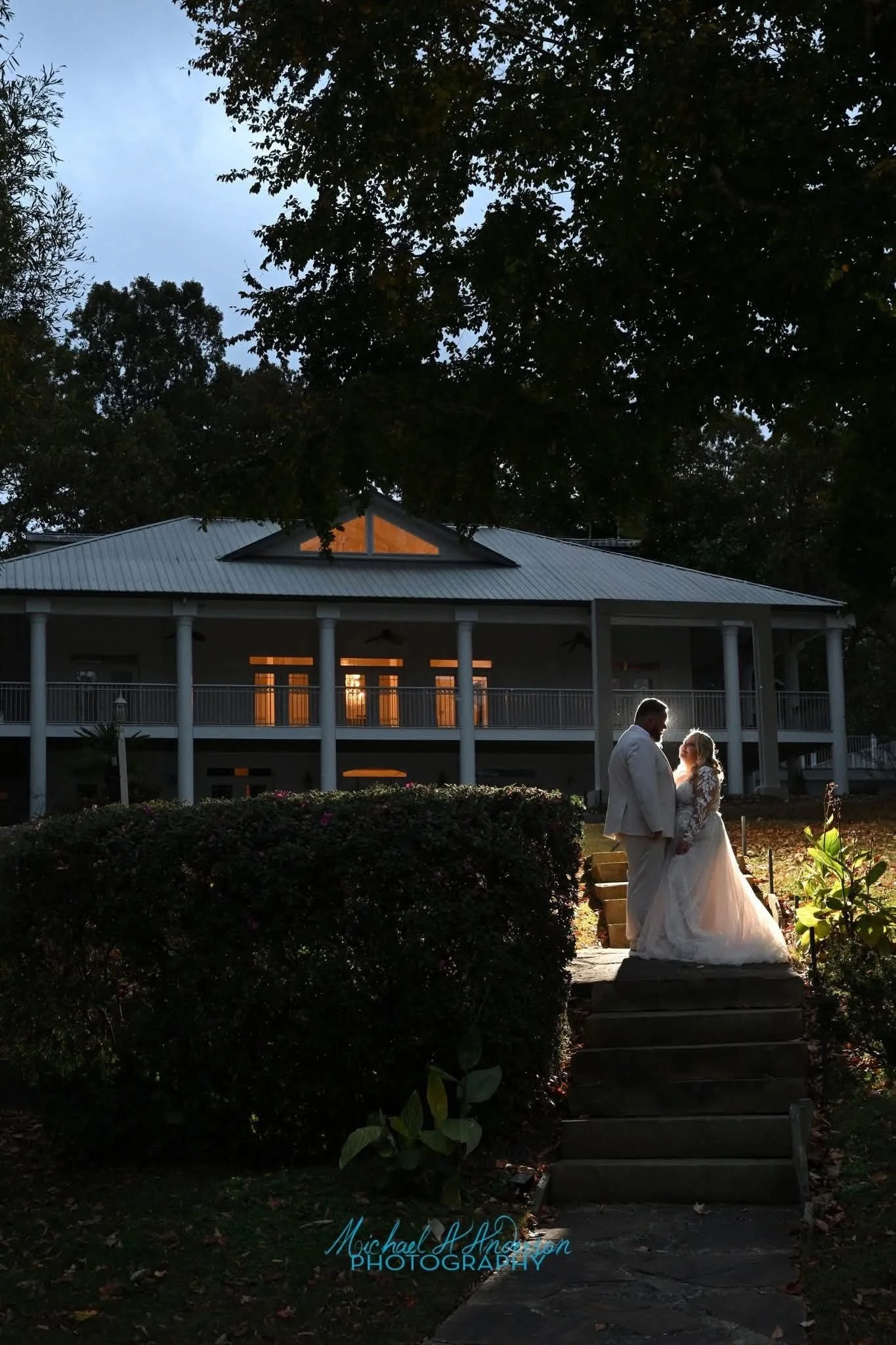 A wedding couple stands on a stone pathway, surrounded by greenery, in front of a large white house with a wrap-around porch, during sunset.