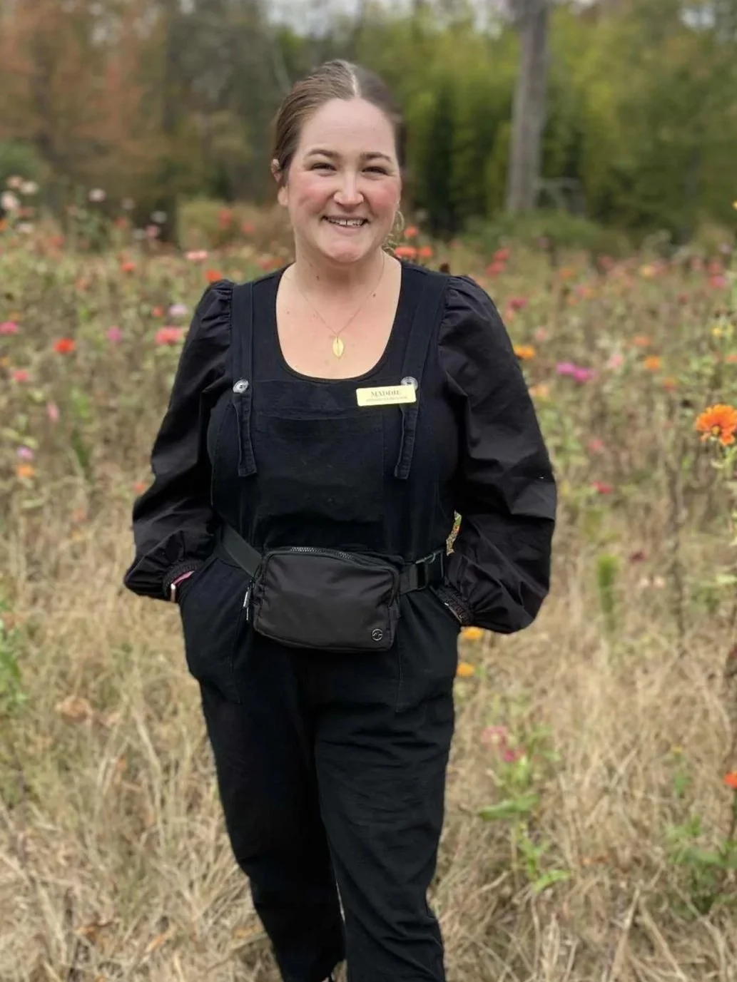 A woman smiling outdoors in a field of flowers and trees, wearing black clothing and a name tag that says "Madeline."