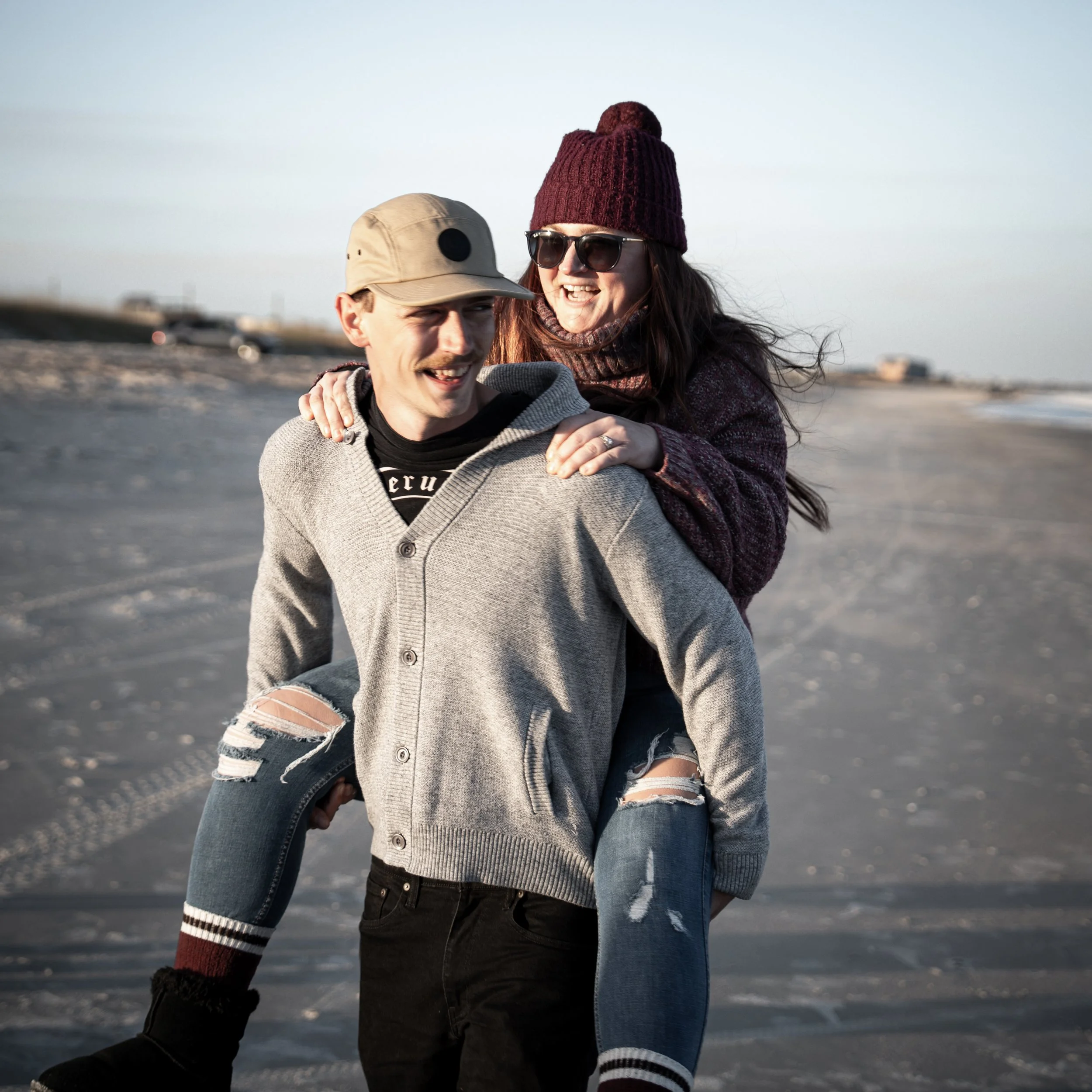 A young woman in a burgundy knit beanie, sunglasses, and a scarf is smiling and leaning on a young man on a beach or lakeside with a clear sky in the background. The young man is wearing a beige cap and a gray hoodie with ripped jeans showing red and white socks, and is smiling.