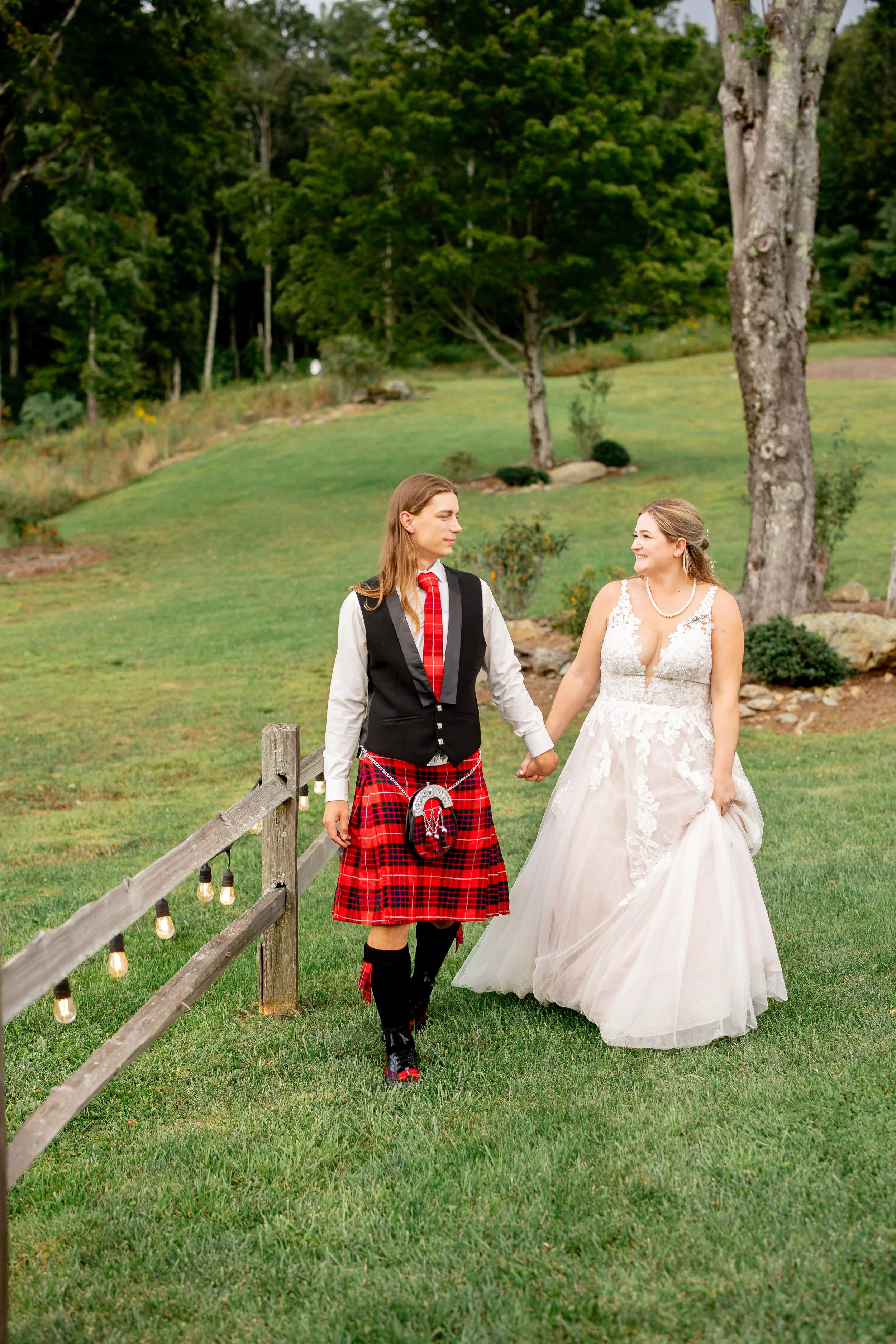 A couple celebrating their wedding walk hand in hand outdoors on a green lawn, with a man dressed in Scottish attire including a kilt, and a woman in a wedding dress, smiling at each other and holding hands.