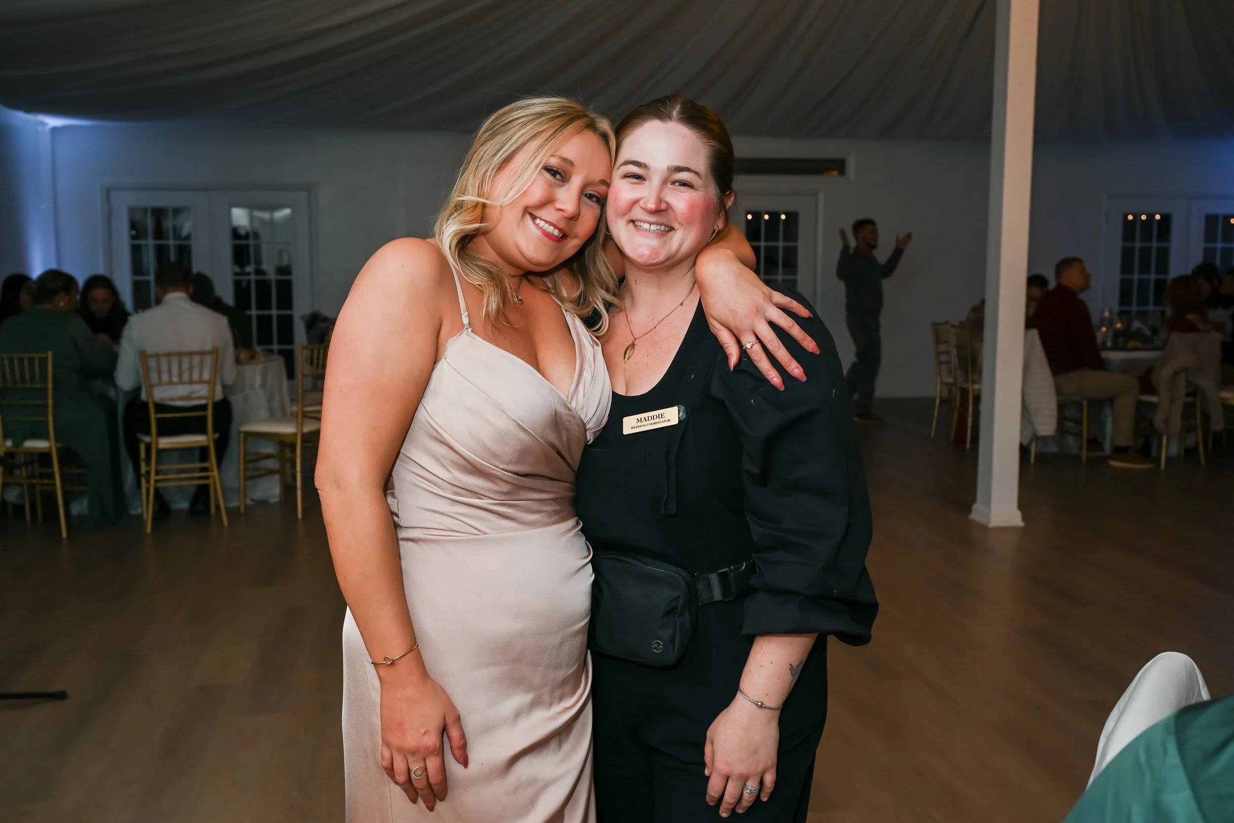 Two women smiling and hugging at an indoor celebration or event, seated at tables with guests in the background.