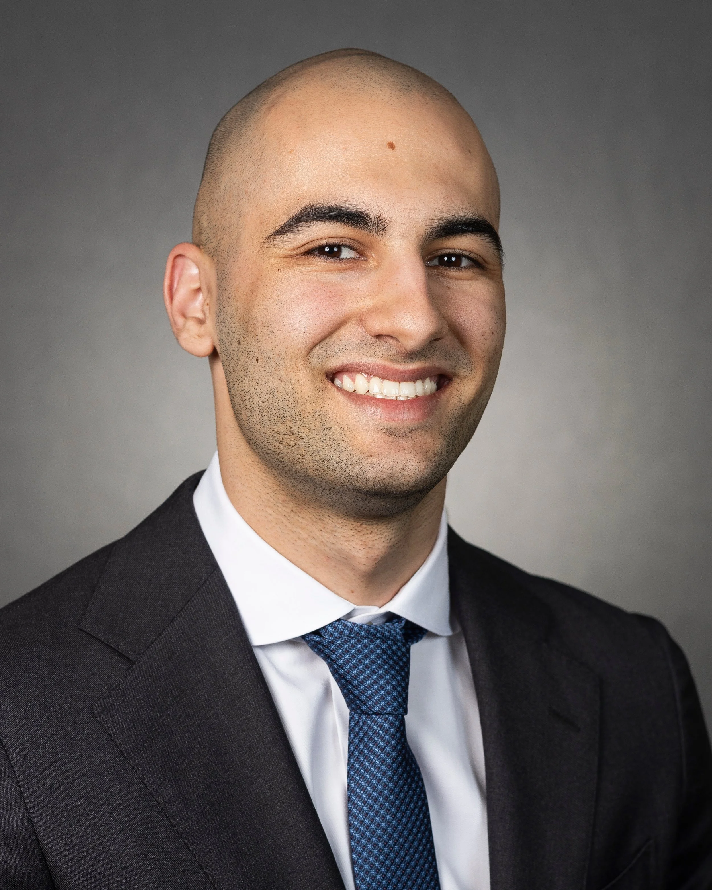 Close-up of a young man with a shaved head wearing a dark suit, white shirt, and blue tie, smiling against a grey background.