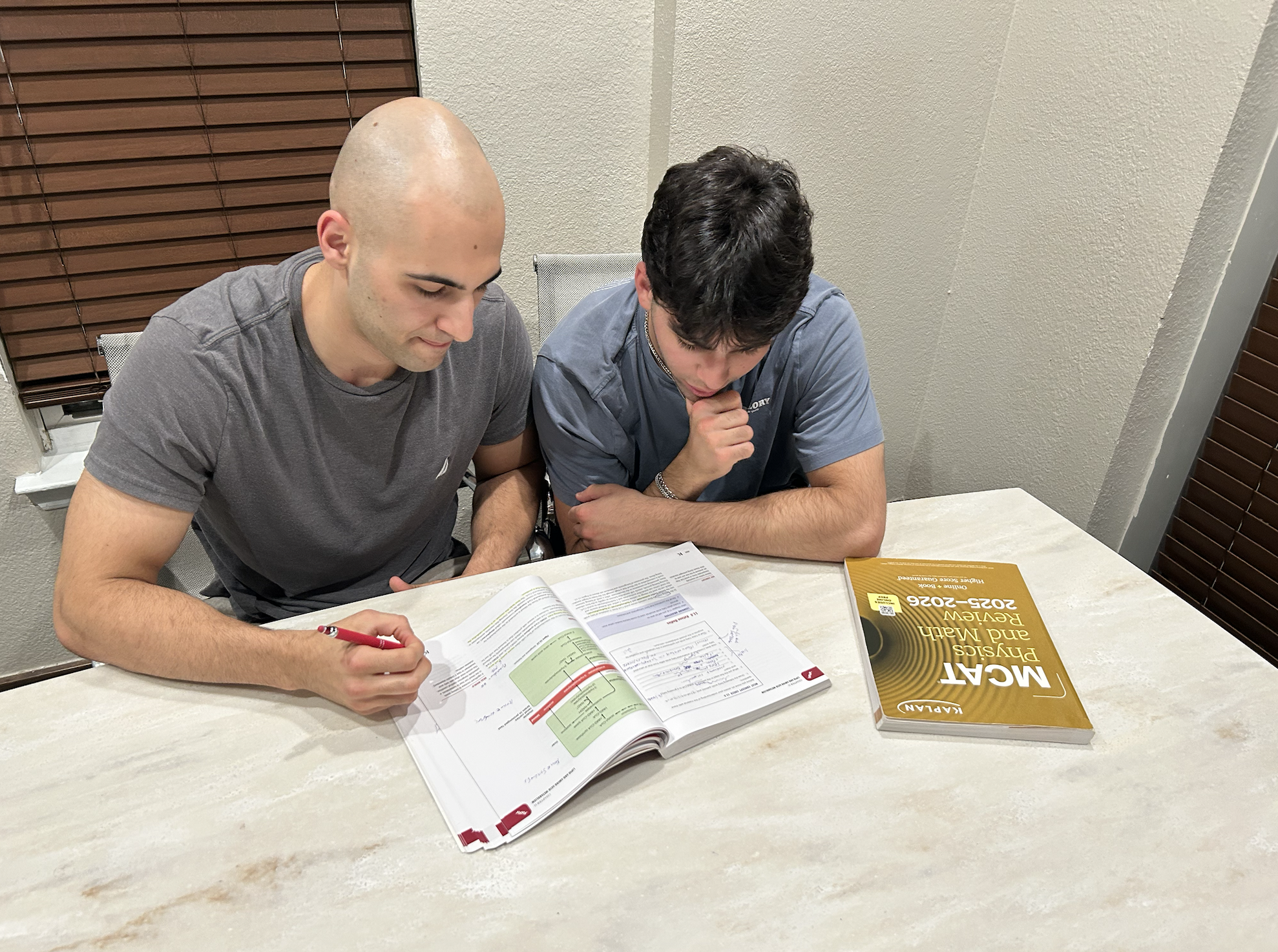 Two young men are seated at a table, studying with open textbooks and notes in front of them. One man has a shaved head and is wearing a gray t-shirt, while the other has dark, wavy hair and is dressed in a blue t-shirt. The setting appears to be a room with beige walls, wooden blinds, and a white table. A recognizable yellow book on the 2025-2026 edition of an academic subject is placed on the table.