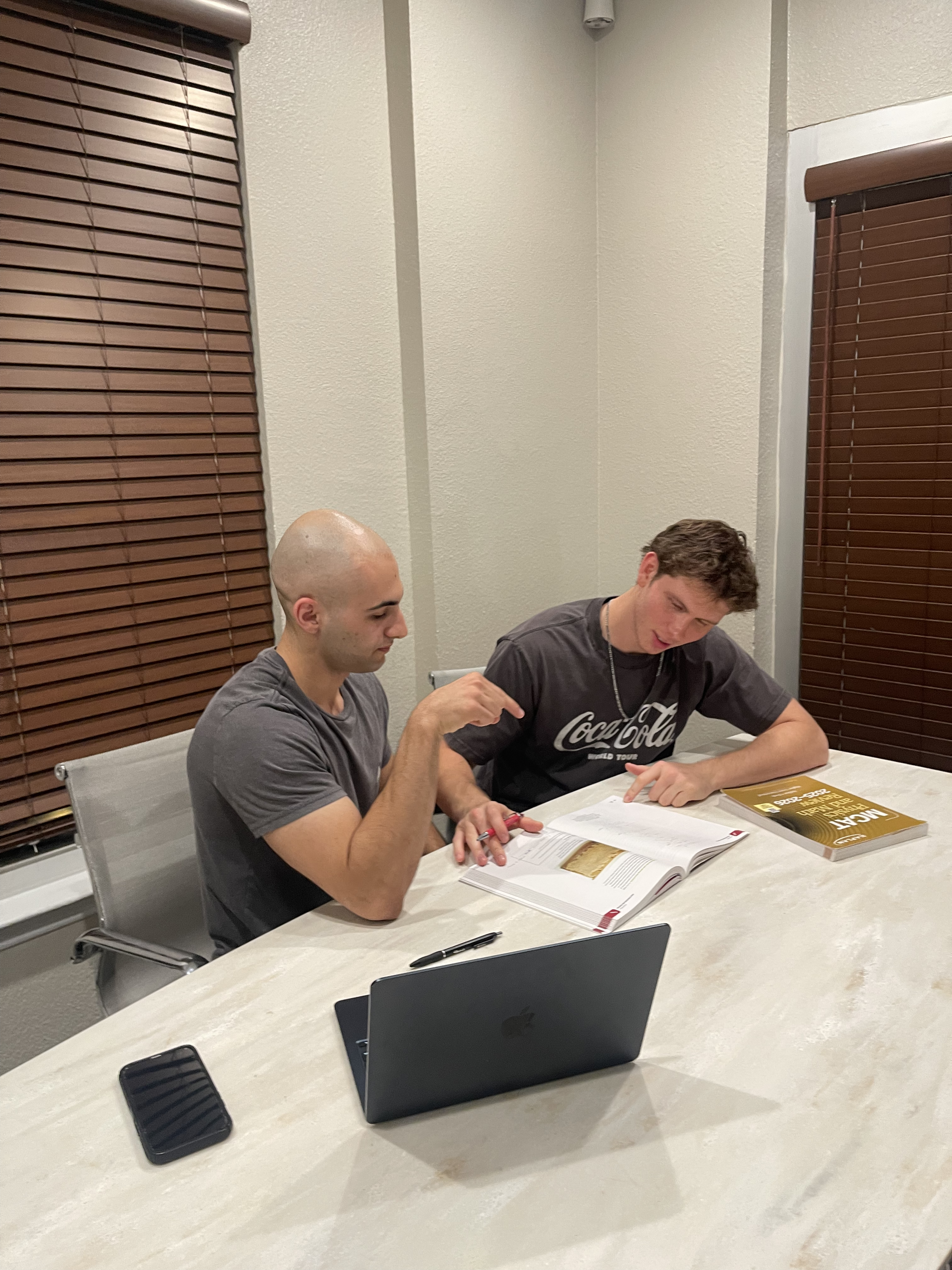 Two young men sitting at a table with open books, a pen, a laptop, and a smartphone, engaged in studying or discussion in a room with beige walls, wooden blinds, and a white table.