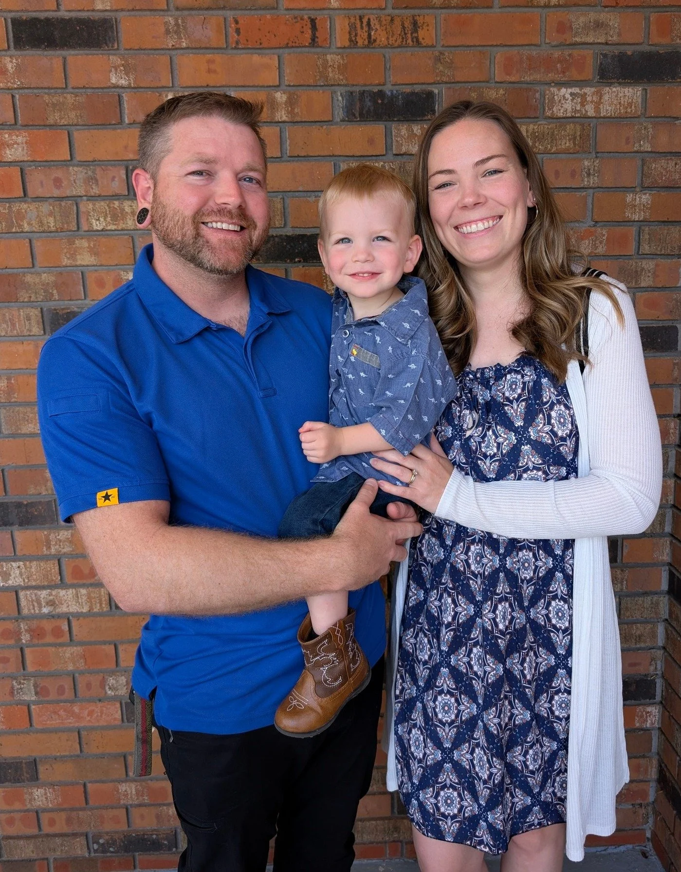 A happy family of three posing in front of a brick wall. The man is holding a young boy, and a woman stands beside them, all smiling.
