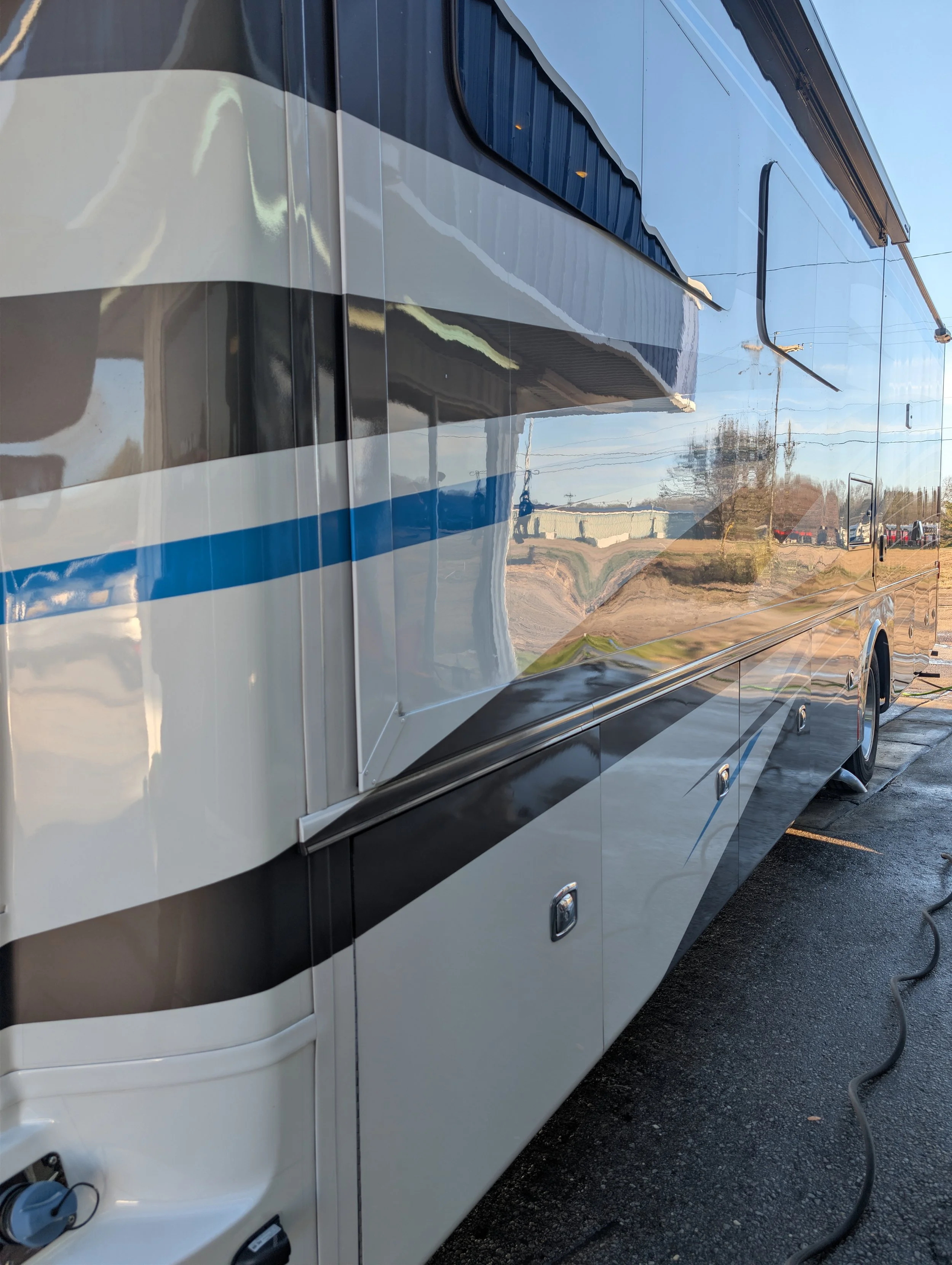 Reflective exterior of a large motorhome parked on a paved lot, with visible power cords and black and white stripes, reflecting the sky and surrounding scenery.