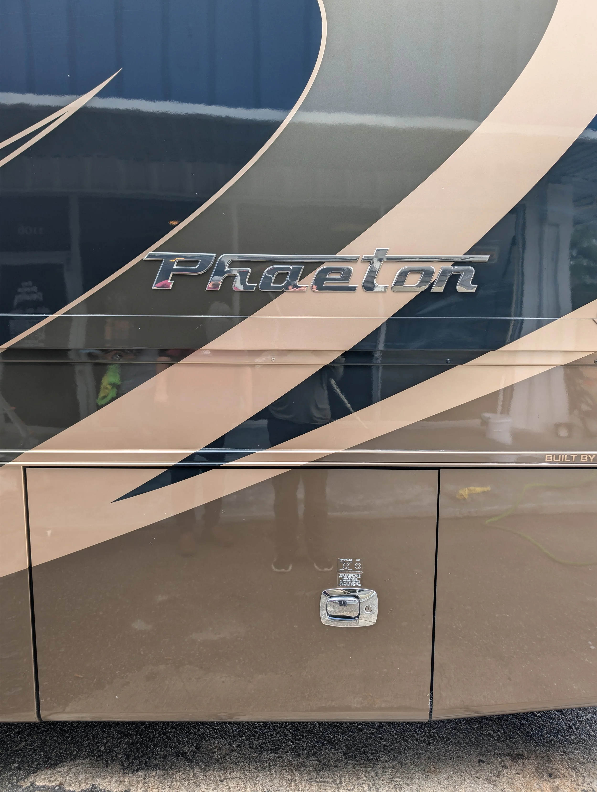 Close-up of the side of a Tiffin Phaeton vehicle with a metallic badge, black and beige graphic design, and a storage compartment with a lock