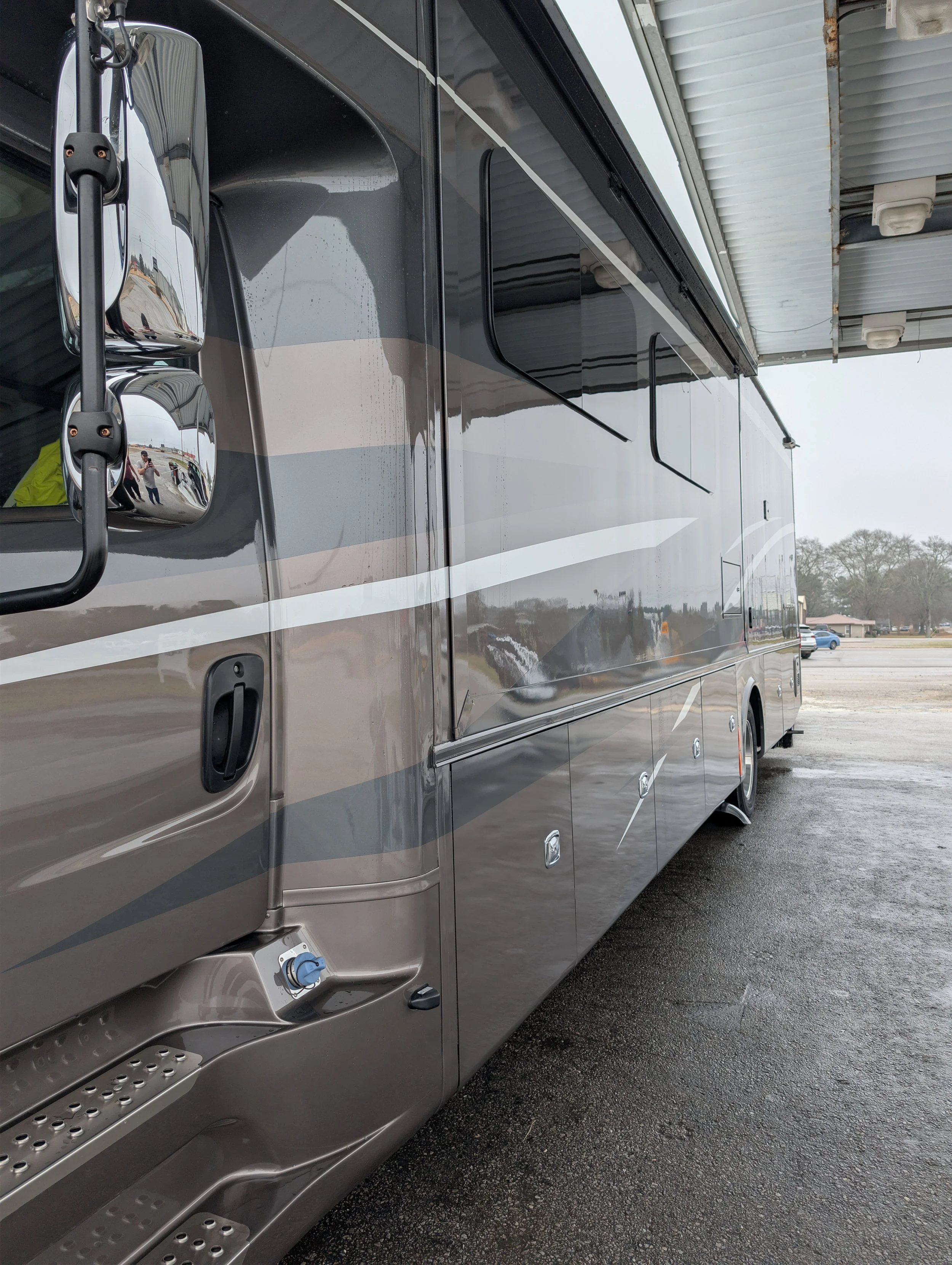 Side view of a large motorhome with reflective exterior, parked under a covered structure on a paved surface, with a cloudy sky and some distant trees and cars in the background.