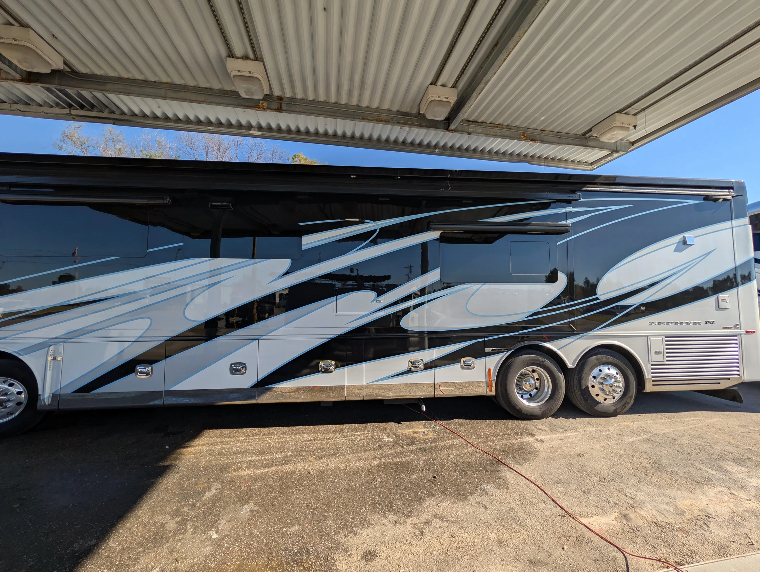 A large black and gray motorhome with blue and white swirl graphics parked outdoors under a metal roof.