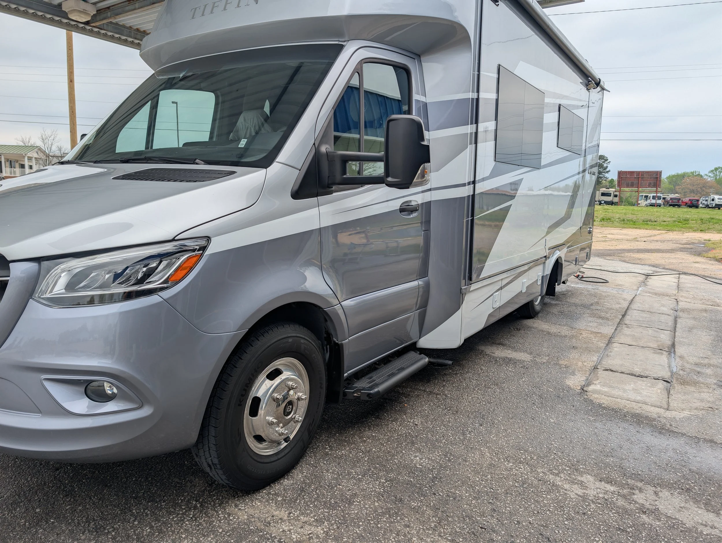 Grey and white Tiffin motorhome parked on a gravel surface with overcast sky in the background.