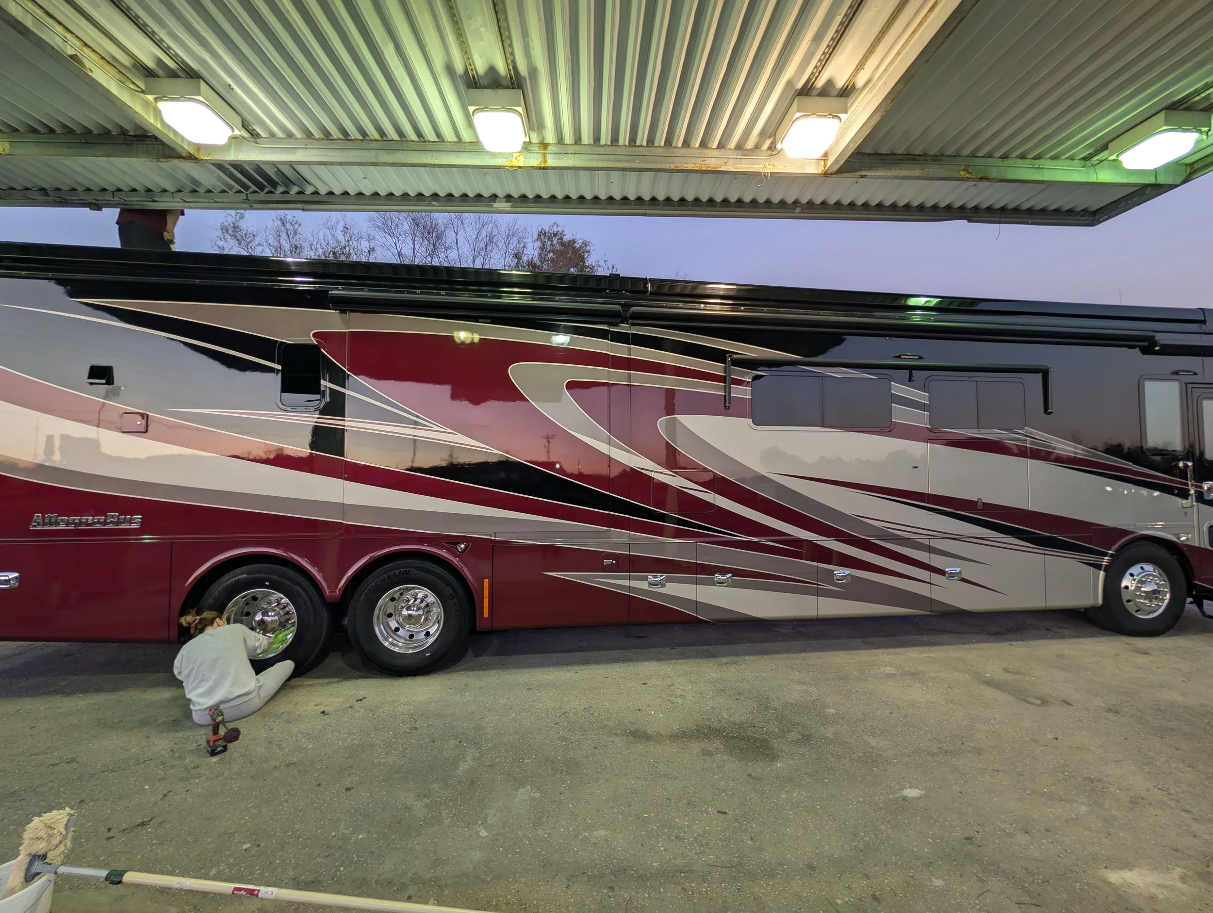 A person working on the rear wheel of a large black, red, and white AllegroBus motorhome parked under a green overhead canopy with bright lights at dusk.