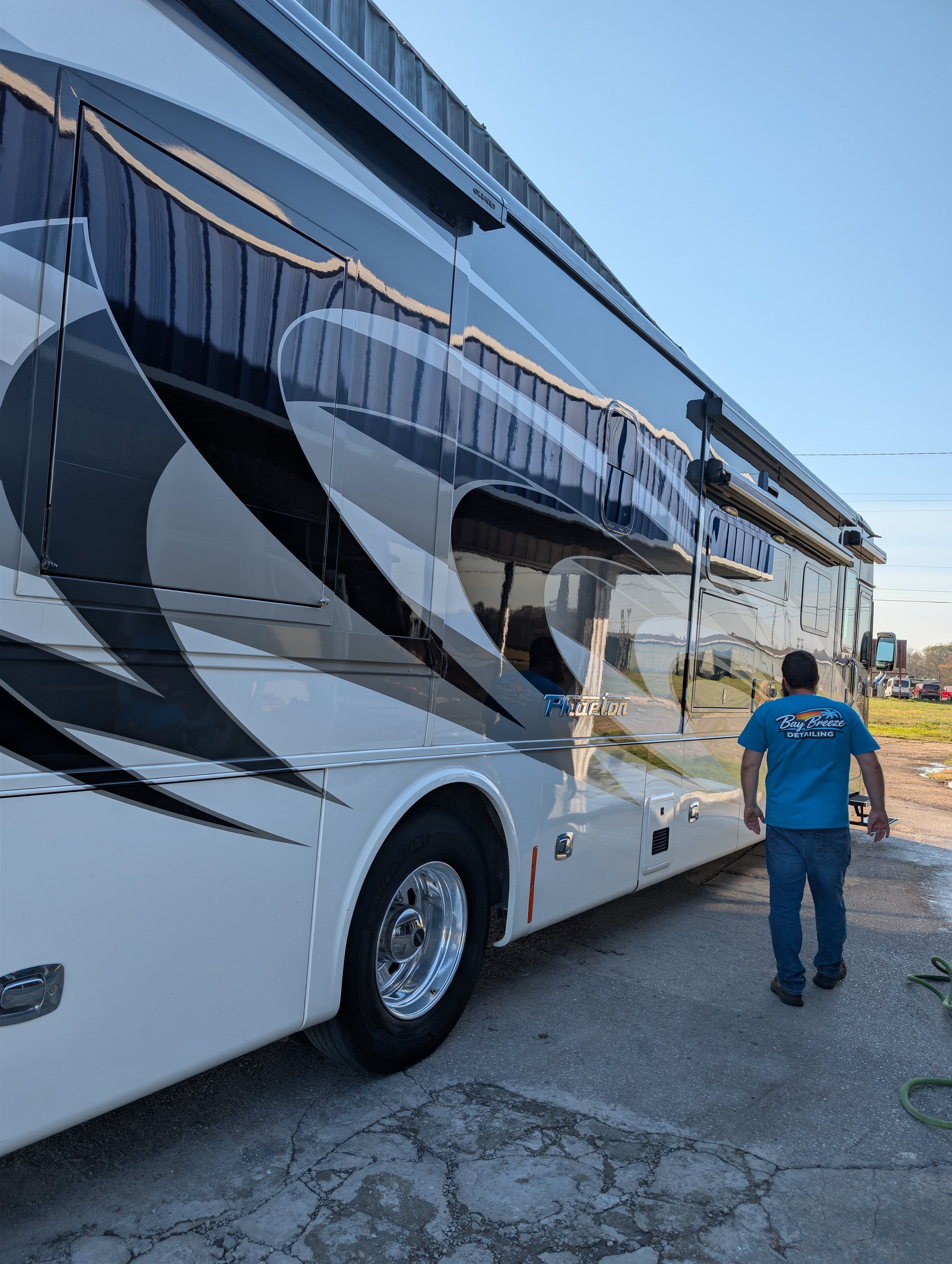 A large, white and black RV with the word "Phaeton" on its side, parked on a cracked pavement with a man in a blue Bay Breeze Detailing shirt walking beside it under a clear blue sky.