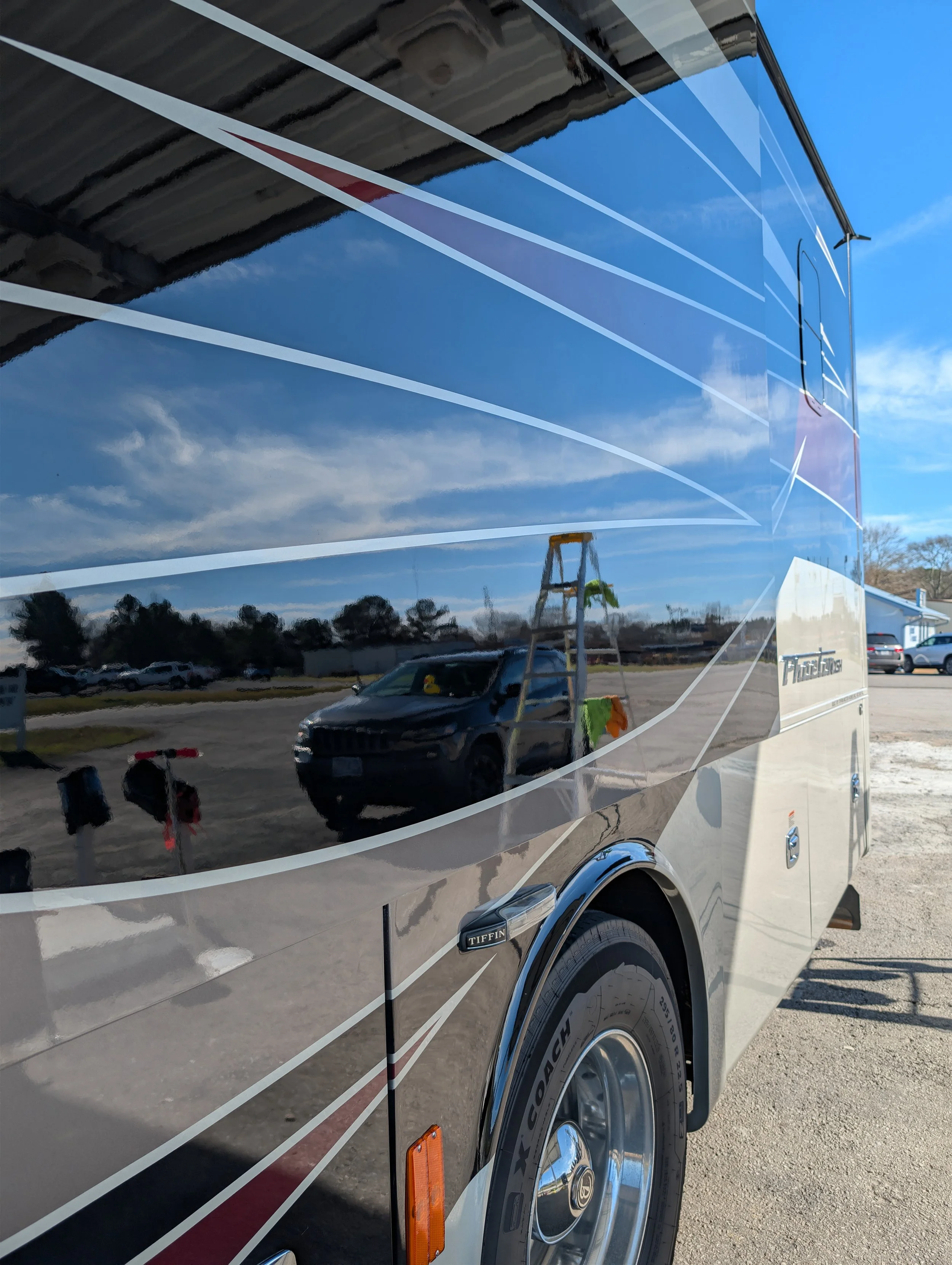 Close-up of a reflective trailer with a shiny surface, showing a reflection of the blue sky with some clouds, parked vehicles, a ladder with cleaning cloths hanging, and a row of mailboxes.