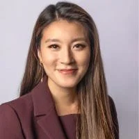 A professional woman with long brown hair, wearing a burgundy blazer and smiling, posed against a plain light gray background.